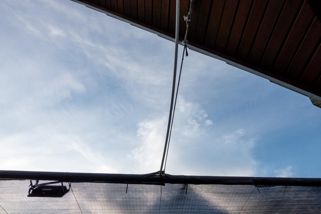Wooden roof with a protruding metal rod extends over a translucent fabric canopy. Blue sky with scattered clouds visible in the background.