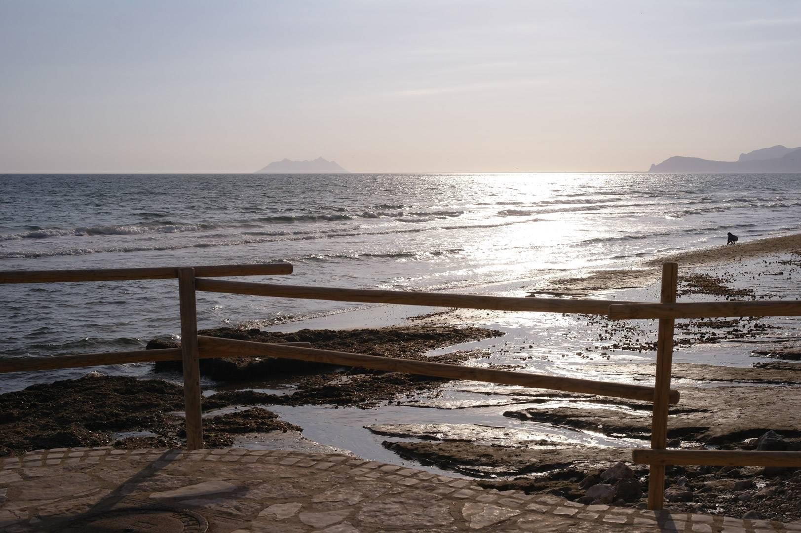 Spiaggia al tramonto, tutta l'immagine ha un colorito terreo che influenza anche il mare. In primo piano, una staccionata di paletti. Sullo sfondo si scorge un'isola, mentre a destra una figura umana accovacciata raccoglie qualcosa dalla sabbia.