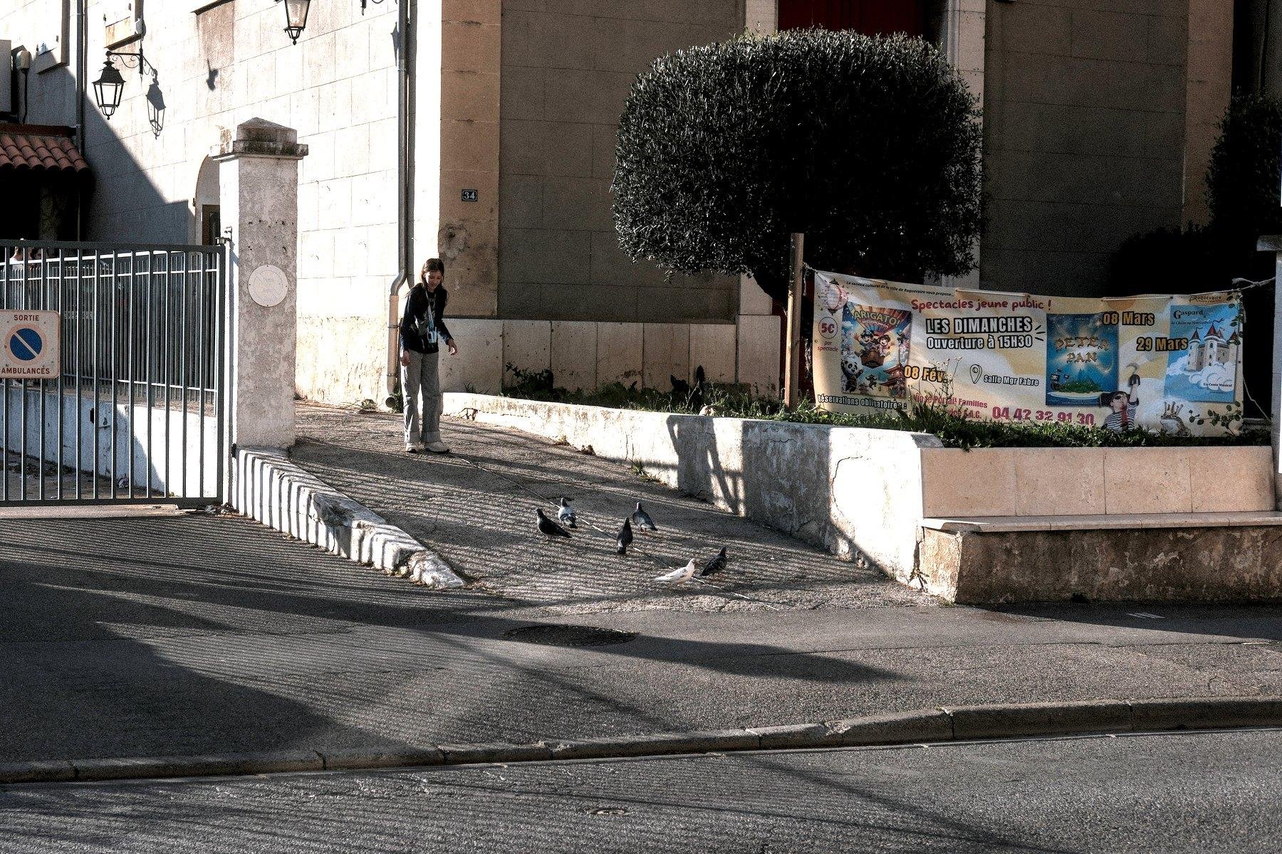 Photo horizontale. Sur un driveway en pente, une jeune fille donne à manger à des pigeons