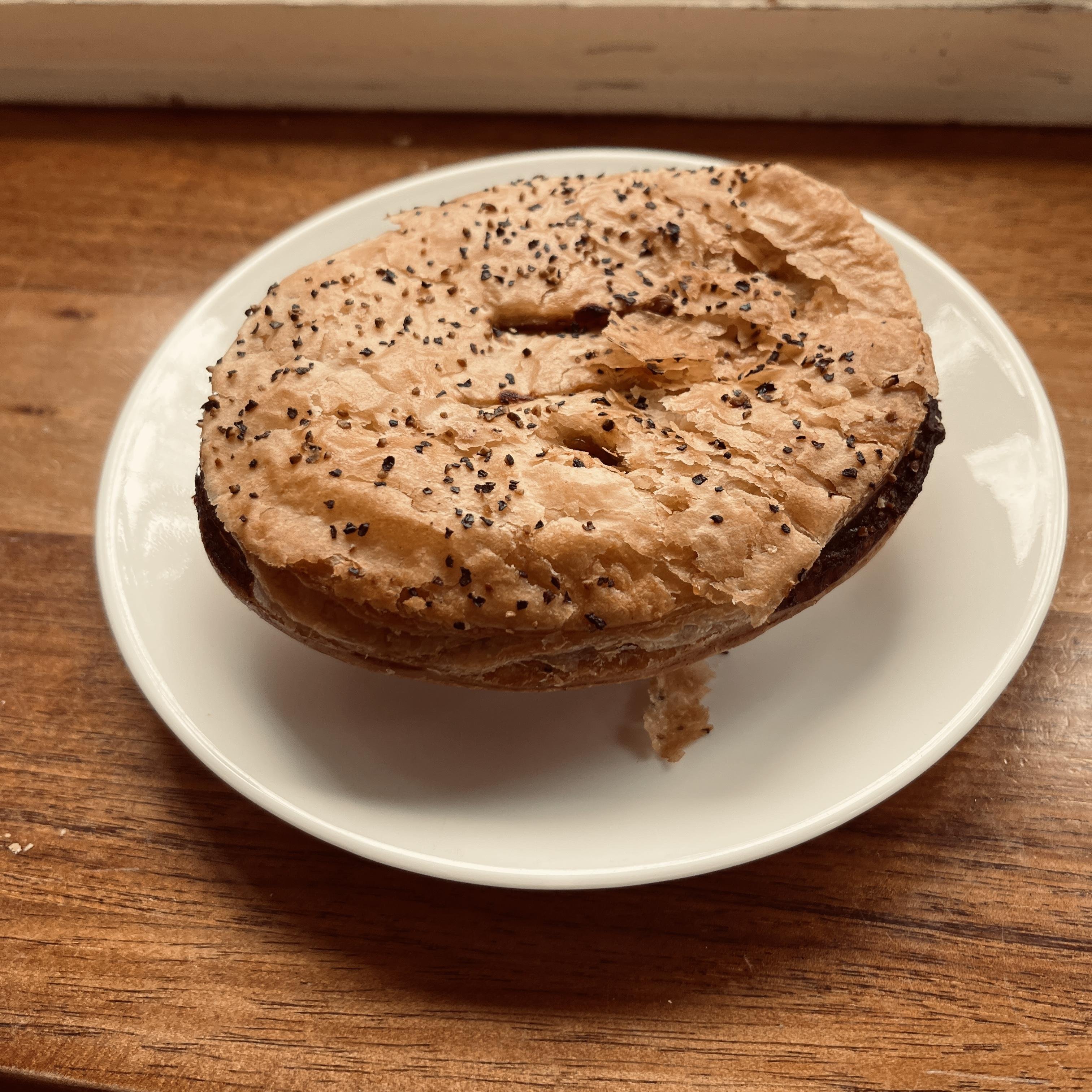 A round, golden-brown poppy seed pepper and steak pie sits on a white plate on a wooden surface. The pie appears slightly toasted with a crisp texture, and poppy seeds are scattered unevenly across the top.