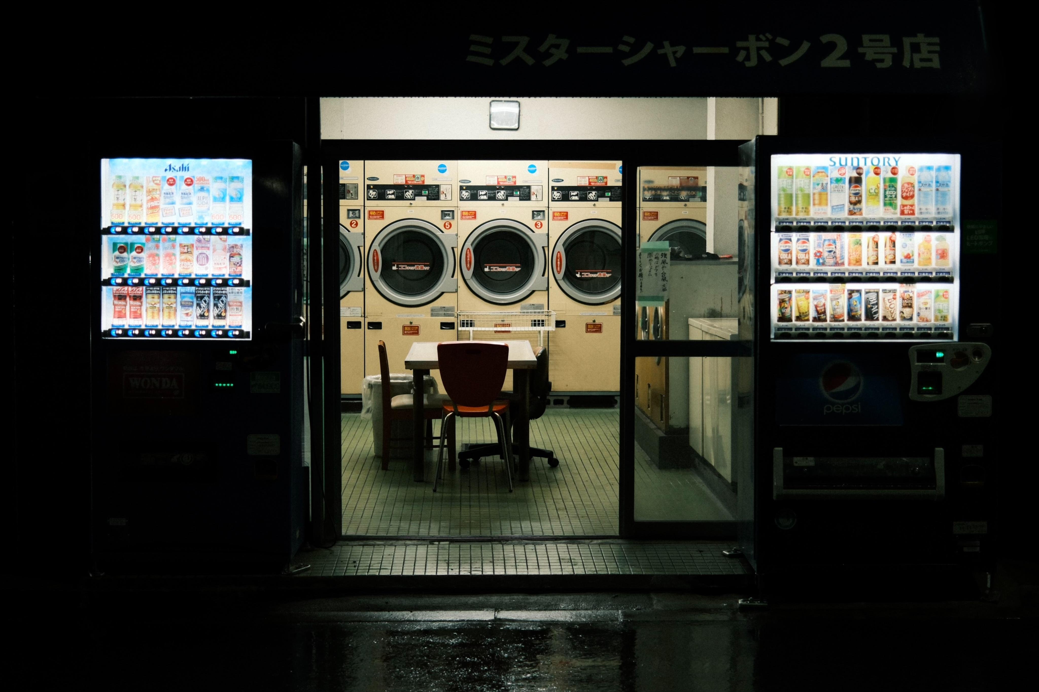 Nighttime view into a laundromat through a glass entrance, with three front-loading washing machines on the back wall and a small table and chairs in the center; illuminated vending machines stand on both sides of the doorway, and wet pavement reflects the lights outside.