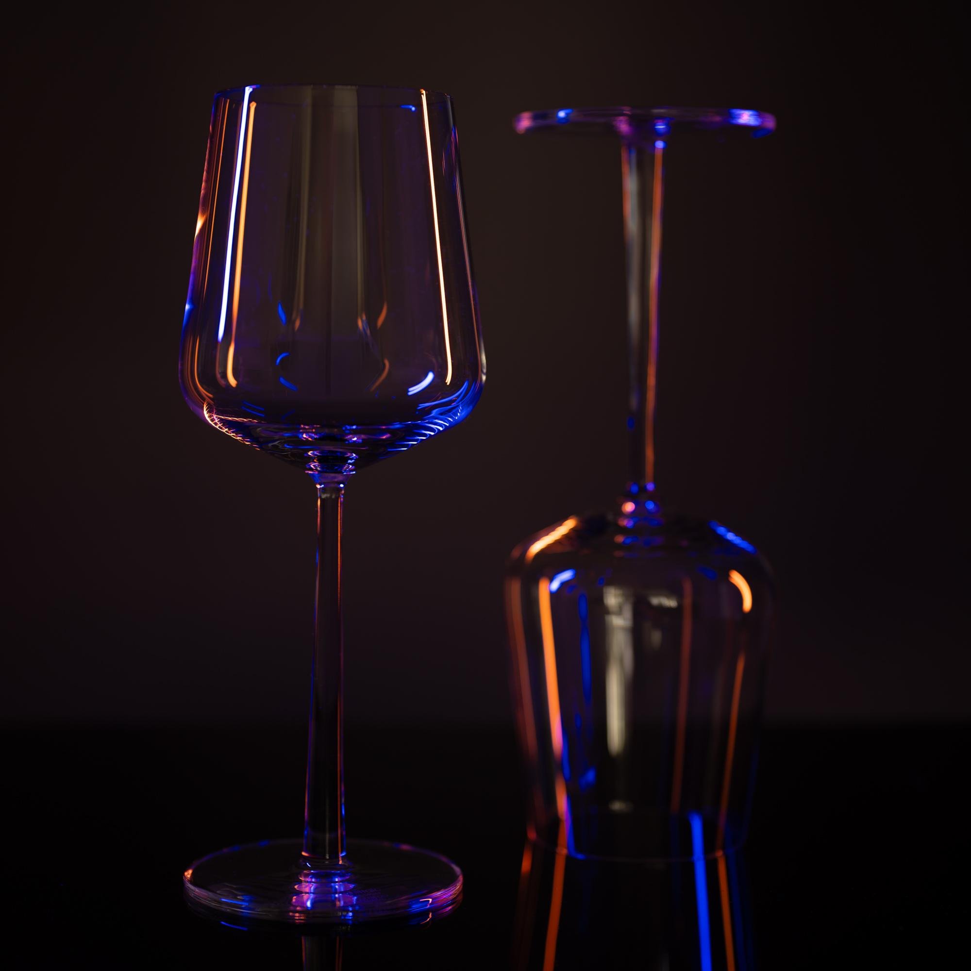The image shows two wine glasses, one upright and the other upside down, on a reflective surface. They are illuminated with colored lighting that creates a mix of blue, purple, and orange reflections on the glass surfaces.
