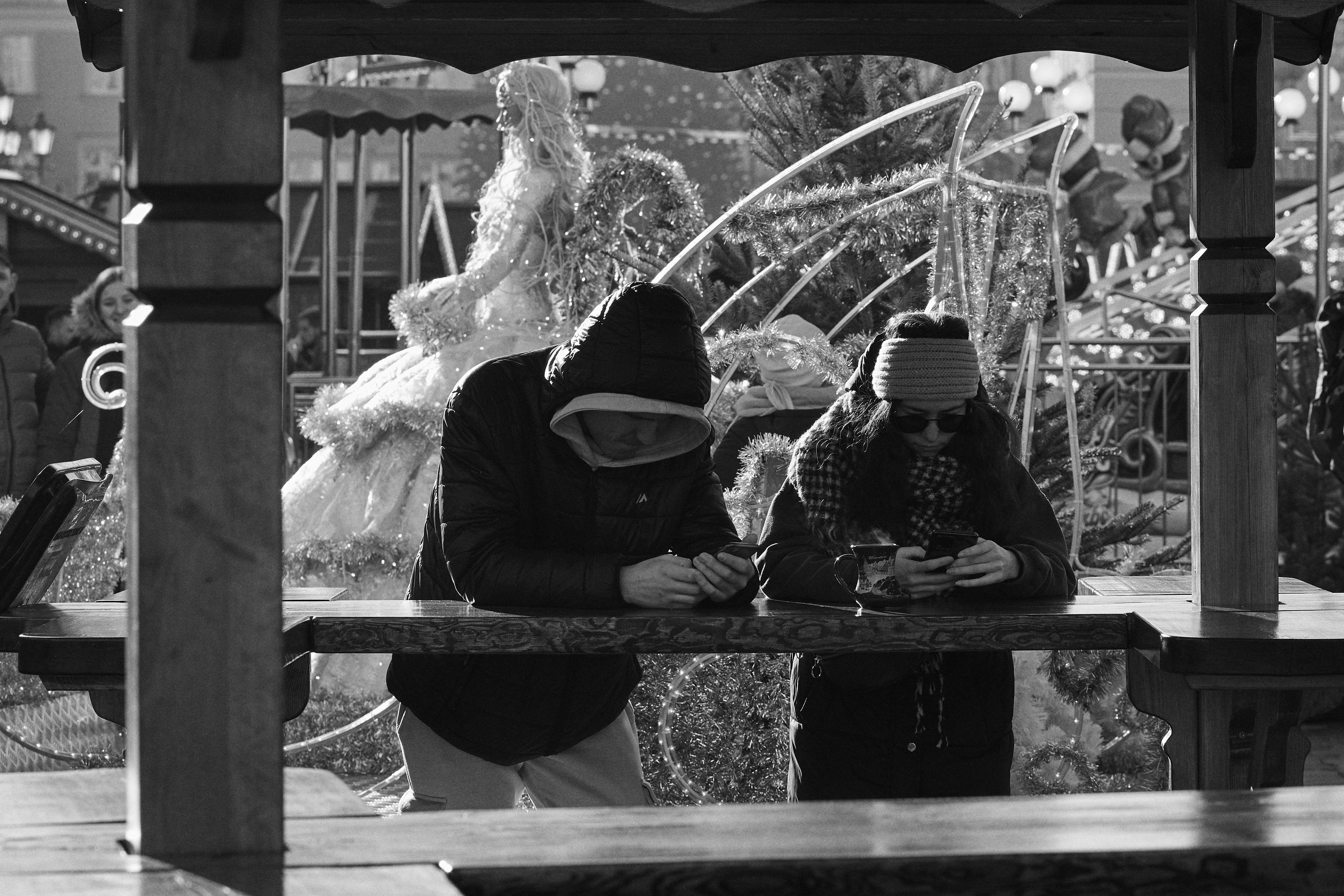 The image depicts two people standing at a wooden table, both engrossed in their phones. They are dressed warmly, suggesting a cold environment. In the background, there's a festive scene with a large, decorative figure resembling an angel or fairy, complete with wings and a gown. The setting appears to be an outdoor event or market.