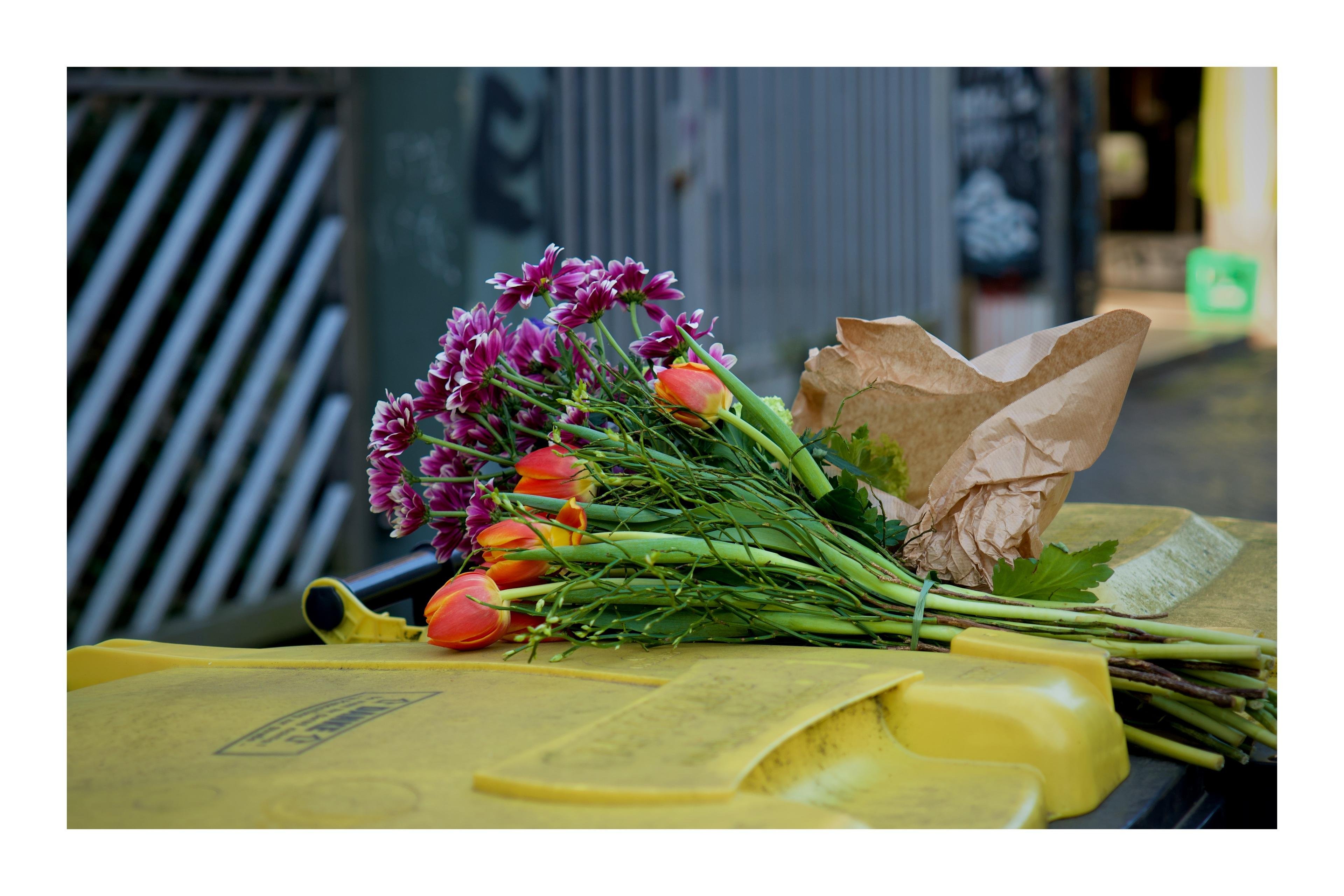 The image shows a bouquet of flowers, including orange tulips and purple daisies, lying on top of a yellow recycling bin. The flowers are partially wrapped in brown paper. 