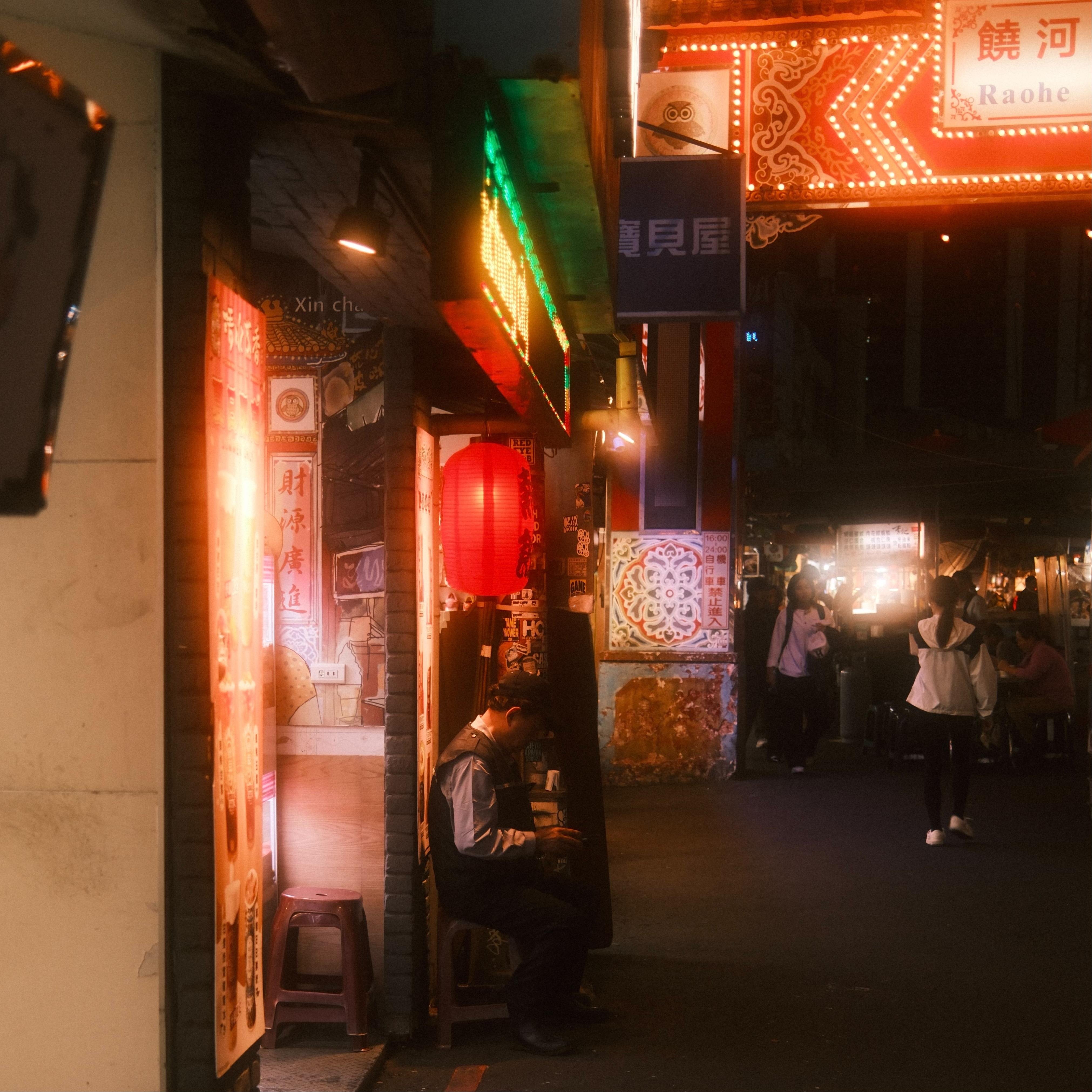 Night street scene with neon signs and Chinese characters above a narrow sidewalk; a red lantern hangs over a doorway where a person sits on a low stool, while pedestrians walk in the dimly lit street to the right.