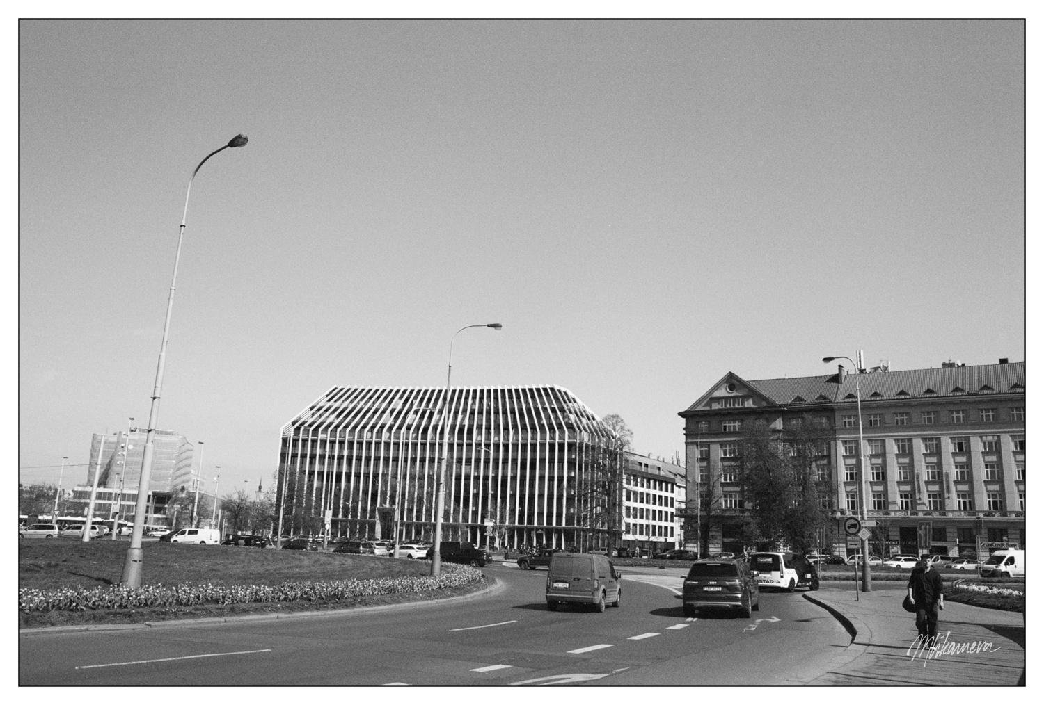 Street scene in black and white with a roundabout in the foreground. A modern building with a distinctive louvered facade stands on the left, while a more traditional multi-story building is on the right. Cars and a van are visible on the road, along with a pedestrian walking on the sidewalk. Several lampposts and trees are scattered throughout the area.