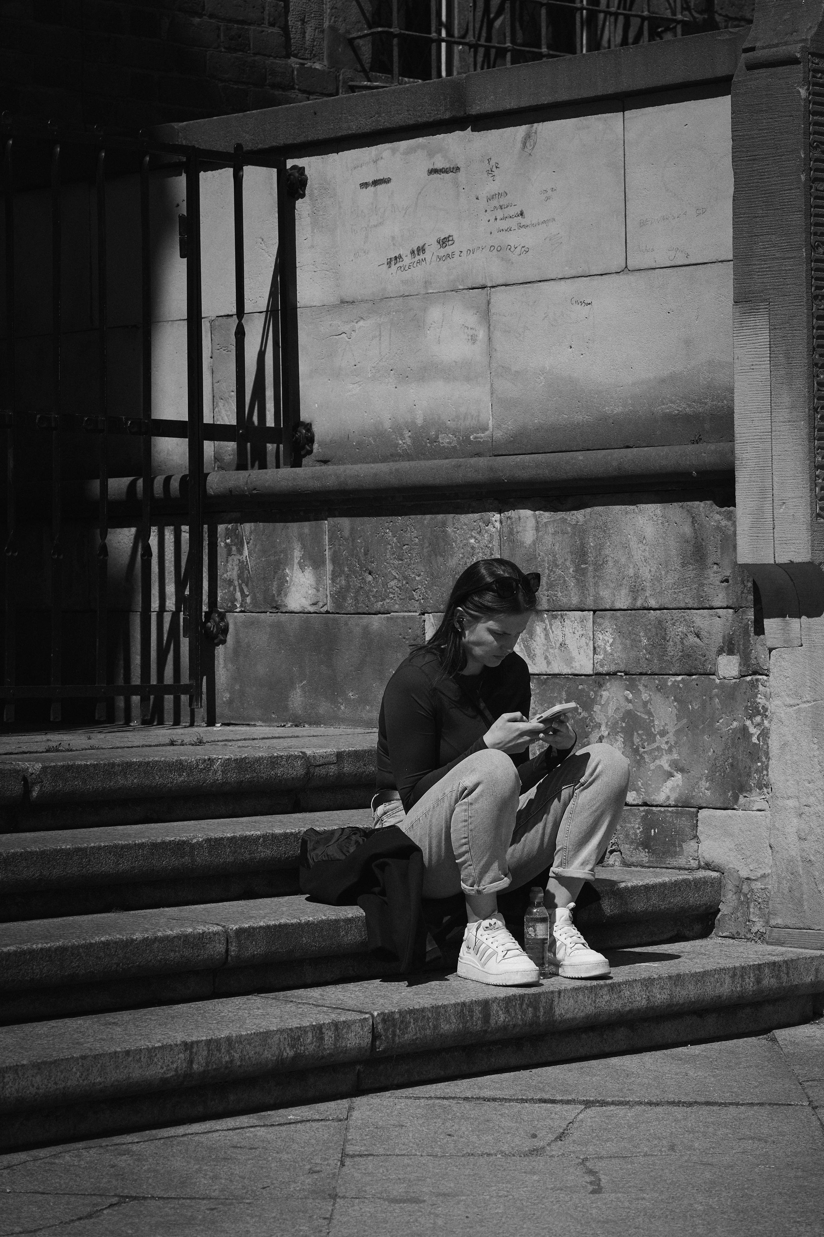 Person sitting on stone steps outside a building, looking down at a smartphone. A jacket and water bottle rest beside them, with a metal gate and large stone wall in the background.