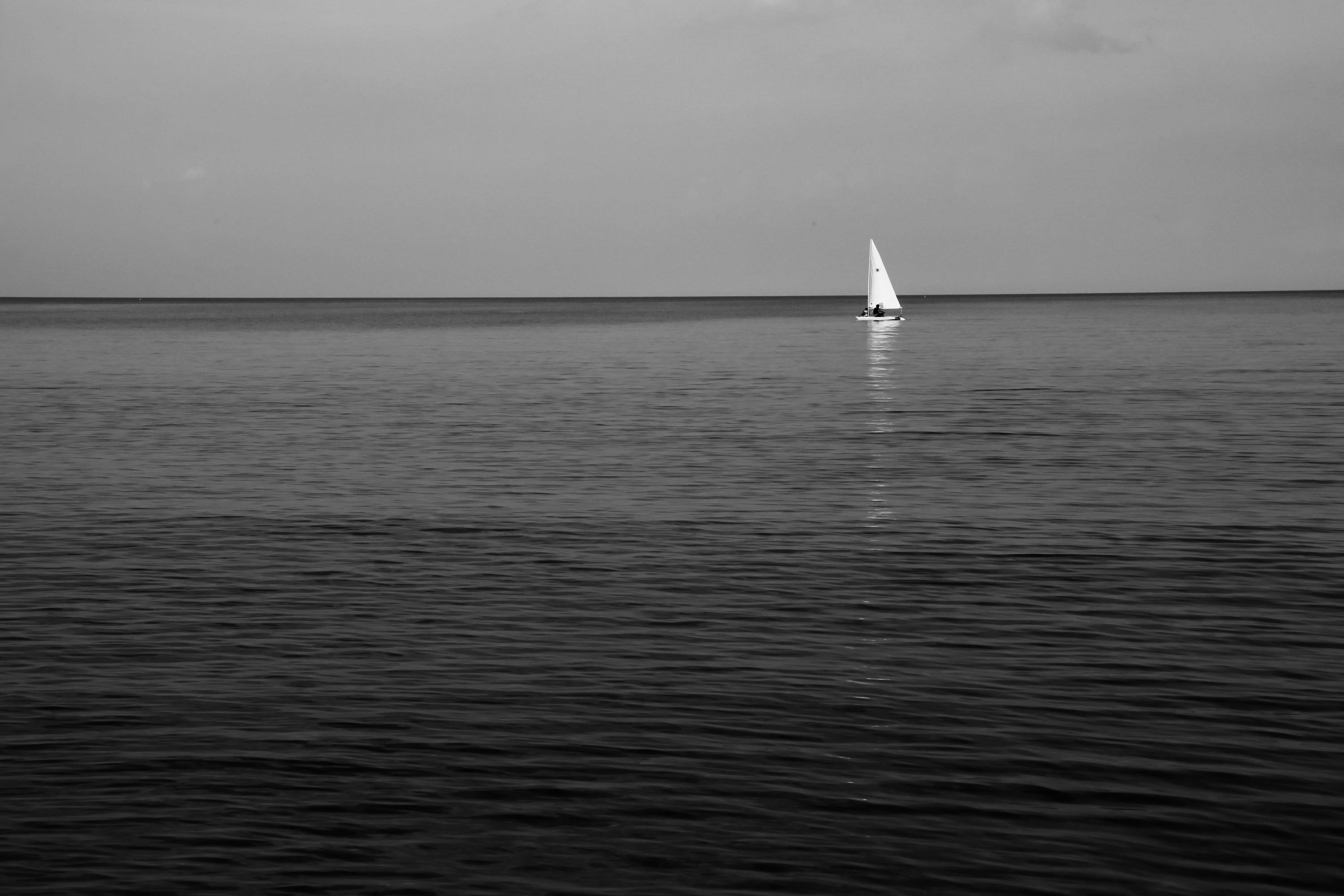 Black-and-white photo of calm ocean water filling most of the frame, a flat horizon across the upper third, and a small sailboat with a white sail near the right side with a faint reflection on the water under an overcast sky.