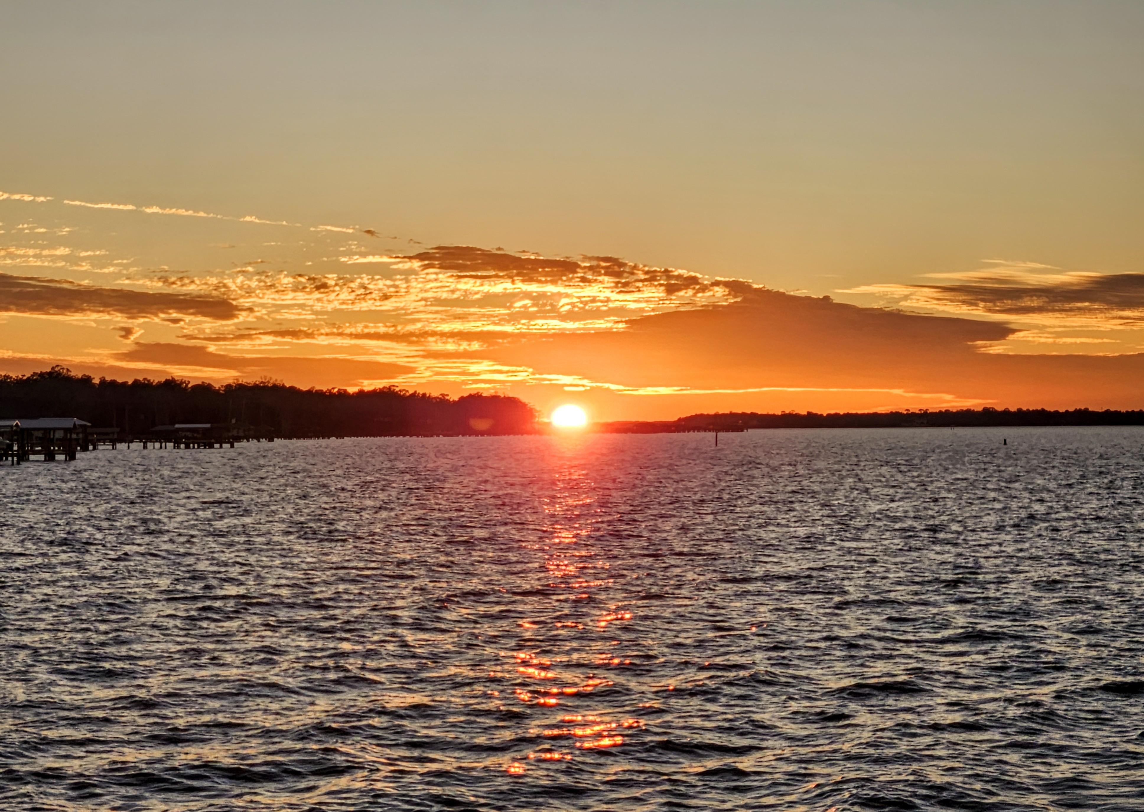 Sunset over a river with rippling waves. The sun is near the horizon, casting an orange glow across the sky and illuminating scattered clouds. A silhouette of a tree line is visible in the distance, and a pier extends into the water on the left side.