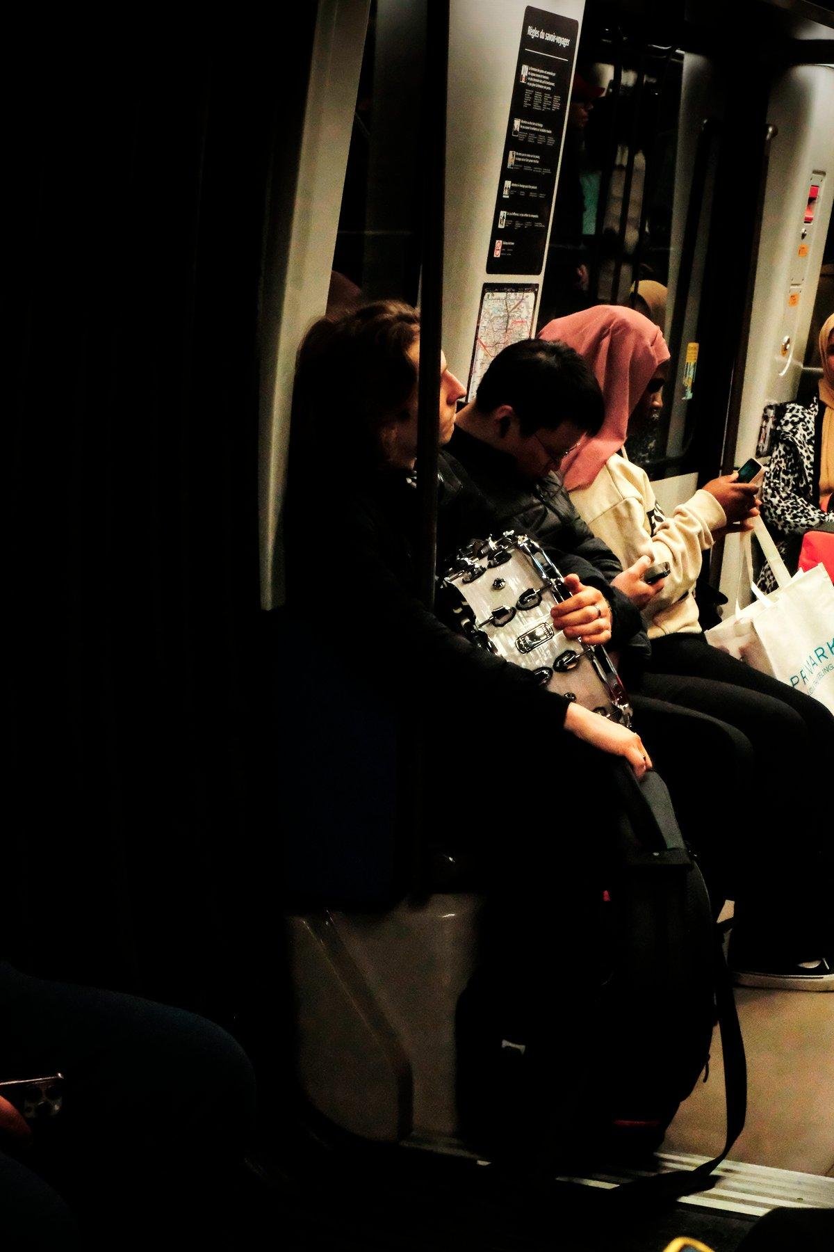 Photo verticale. Dans une rame de métro, Un homme assis tiens contre lui une caisse claire de batterie blanche, et un sac noir qui doit sûrement contenir ses cymbales, vu la forme en disque