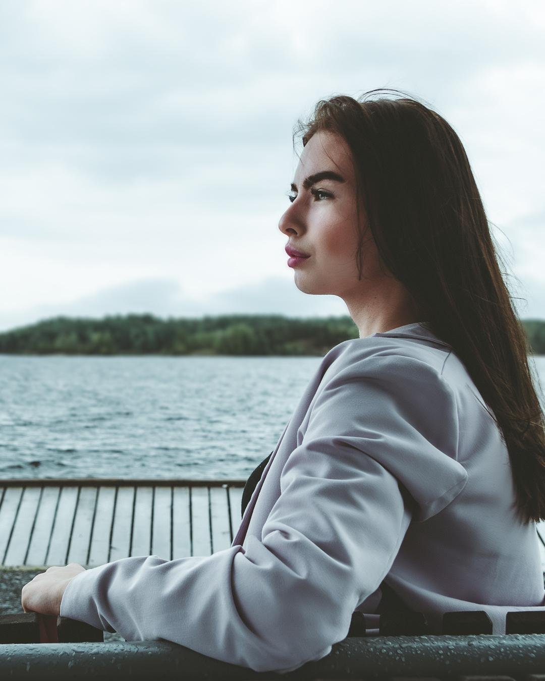 The image shows a person with long dark hair gazing thoughtfully at a body of water. The person is wearing a light-colored jacket and is seated outdoors, with a lake and a wooded area in the background. The scene has a calm and serene atmosphere. Valokuvaaja Kuvaajankulma Lappeenranta