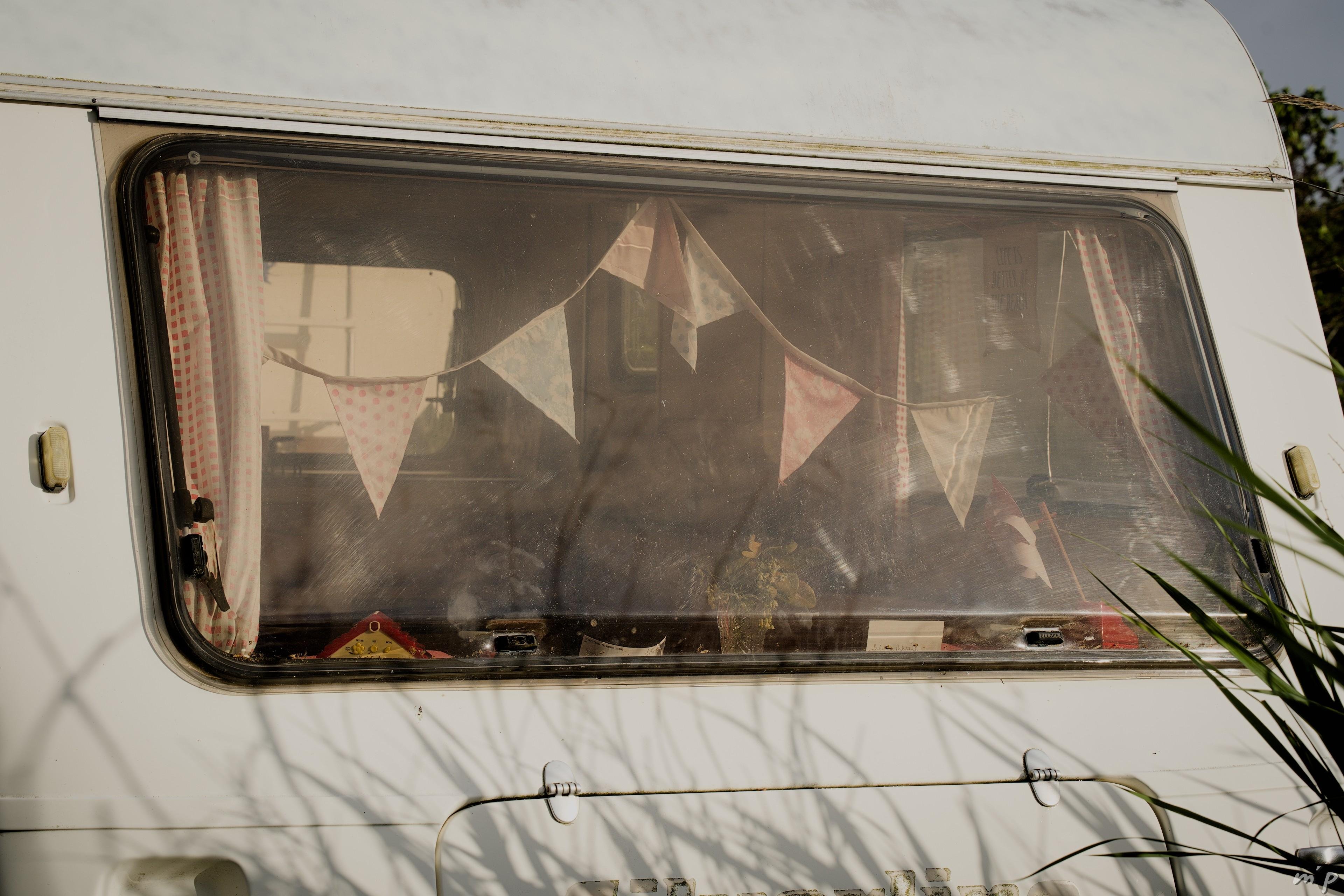 🇬🇧 Horizontal color photo of a caravan window in a garden. A garland of sun-washed pennants hangs behind the window. We can also see the curtains and some trinkets. The image exudes a certain nostalgia.

🇫🇷 Photo couleur horizontale d'une fenêtre de caravane dans un jardin. Une girlande de fanions délavés par le soleil est suspendues derrière la fenêtre. On distingue également les rideaux et quelques babioles. L'image dégage une certain nostalgie.