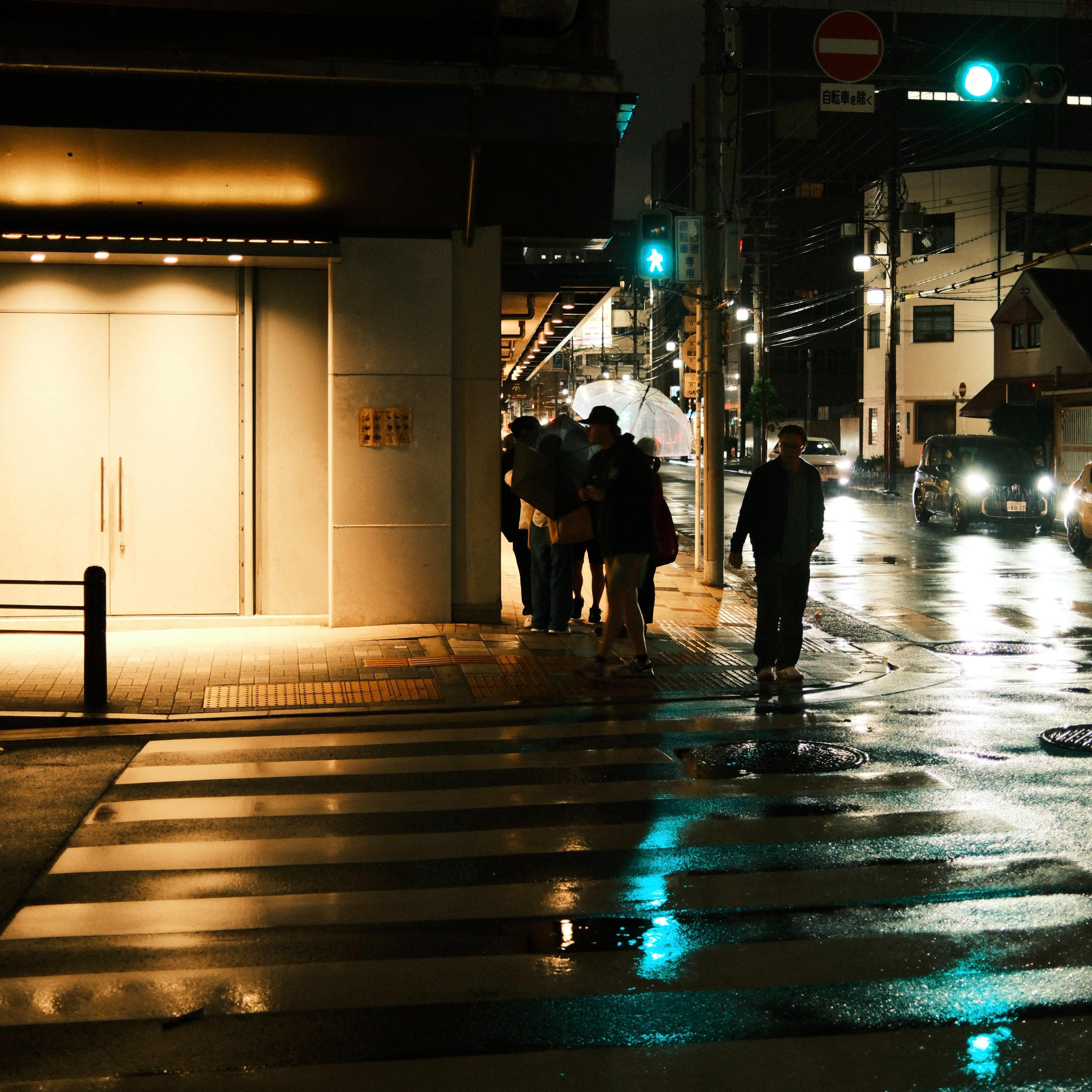 Rainy nighttime street intersection with a zebra crosswalk in the foreground, wet pavement reflecting lights, pedestrians under umbrellas near a building entrance on the left, and cars with headlights on the right beneath green traffic signals and overhead wires.