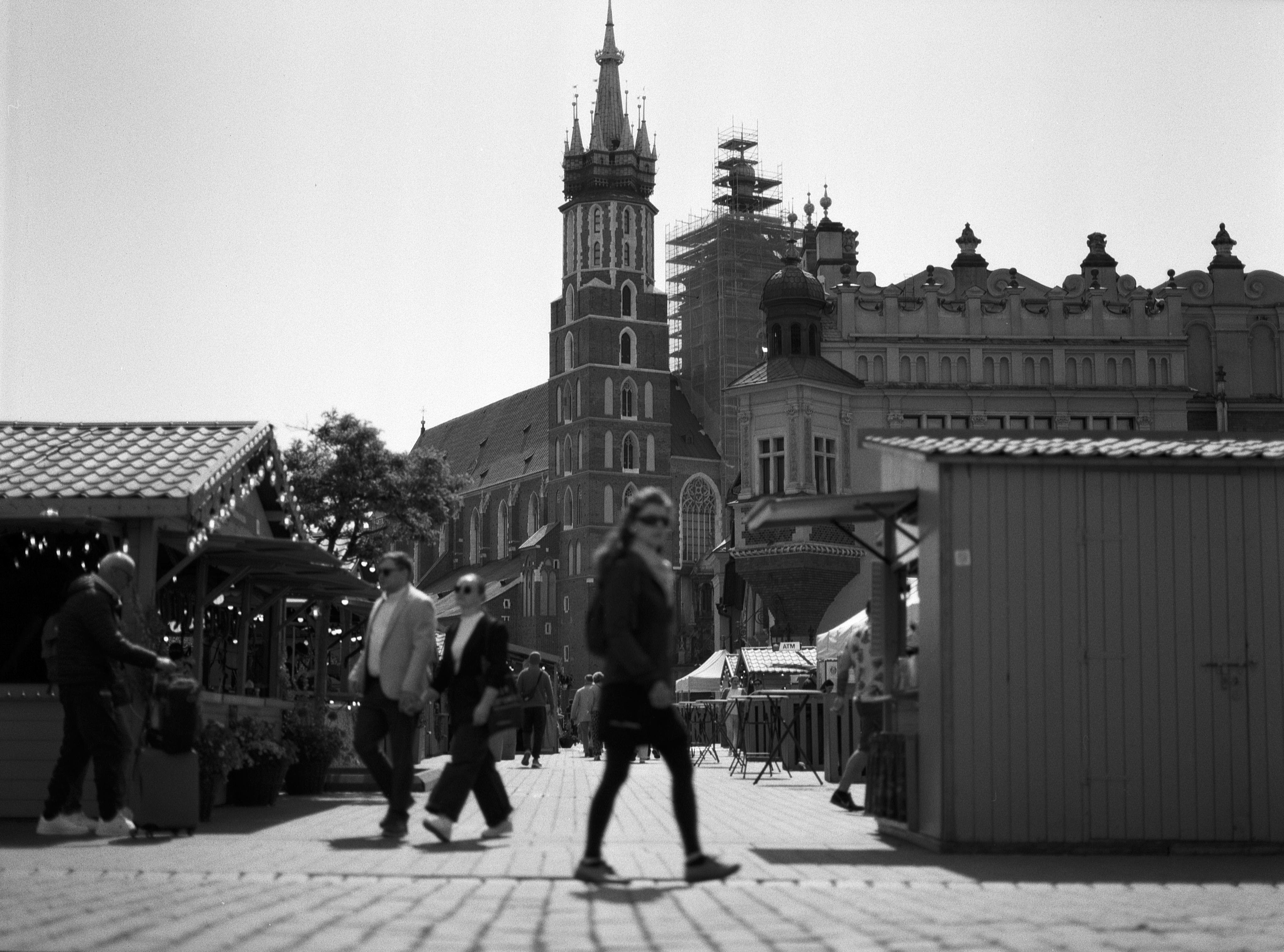 Black and white photo of a bustling square with several people walking. In the background, a large historic church with scaffolding around its tower and ornate architectural details. Wooden market stalls line the square in the foreground. The scene conveys a mix of modern and historical city life.
