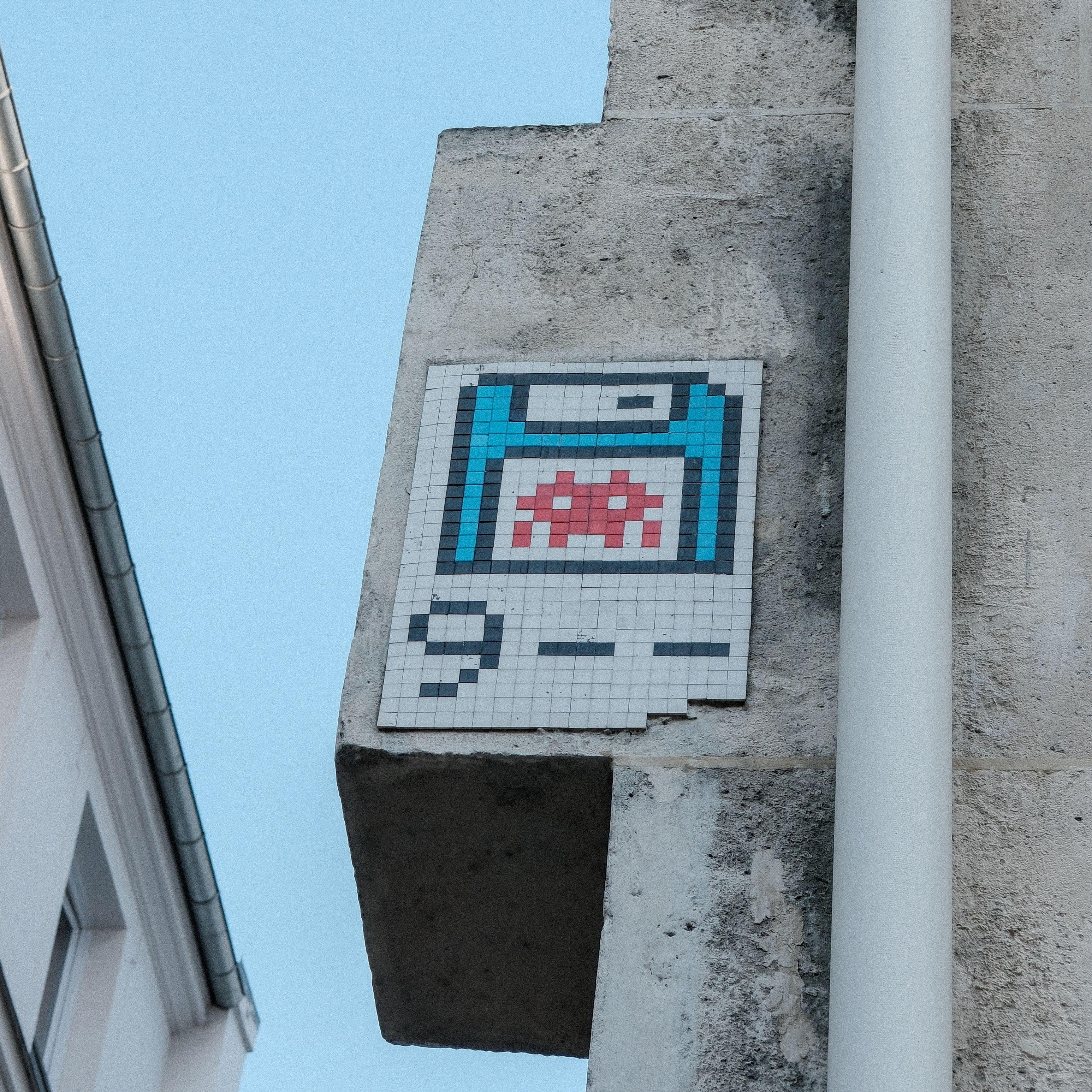 Photo carrée. Sur le mur d’un immeuble, un street art mosaïc représentant une disquette bleu, avec un space invader sur son étiquette. En dessous, on peut lire « 9 - - »