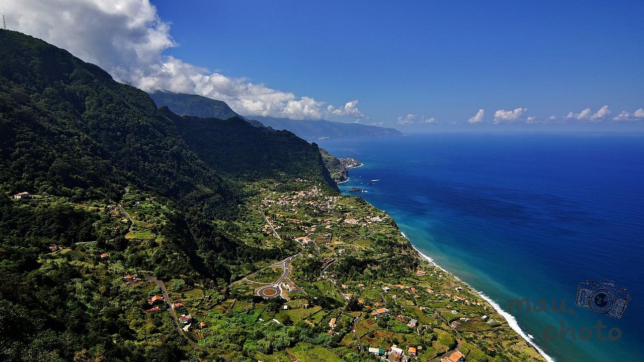 Aerial view of a steep, forested mountain ridge dropping to a coastal plain with scattered houses, fields, winding roads, and a roundabout; a narrow shoreline borders deep blue ocean under a partly cloudy sky.