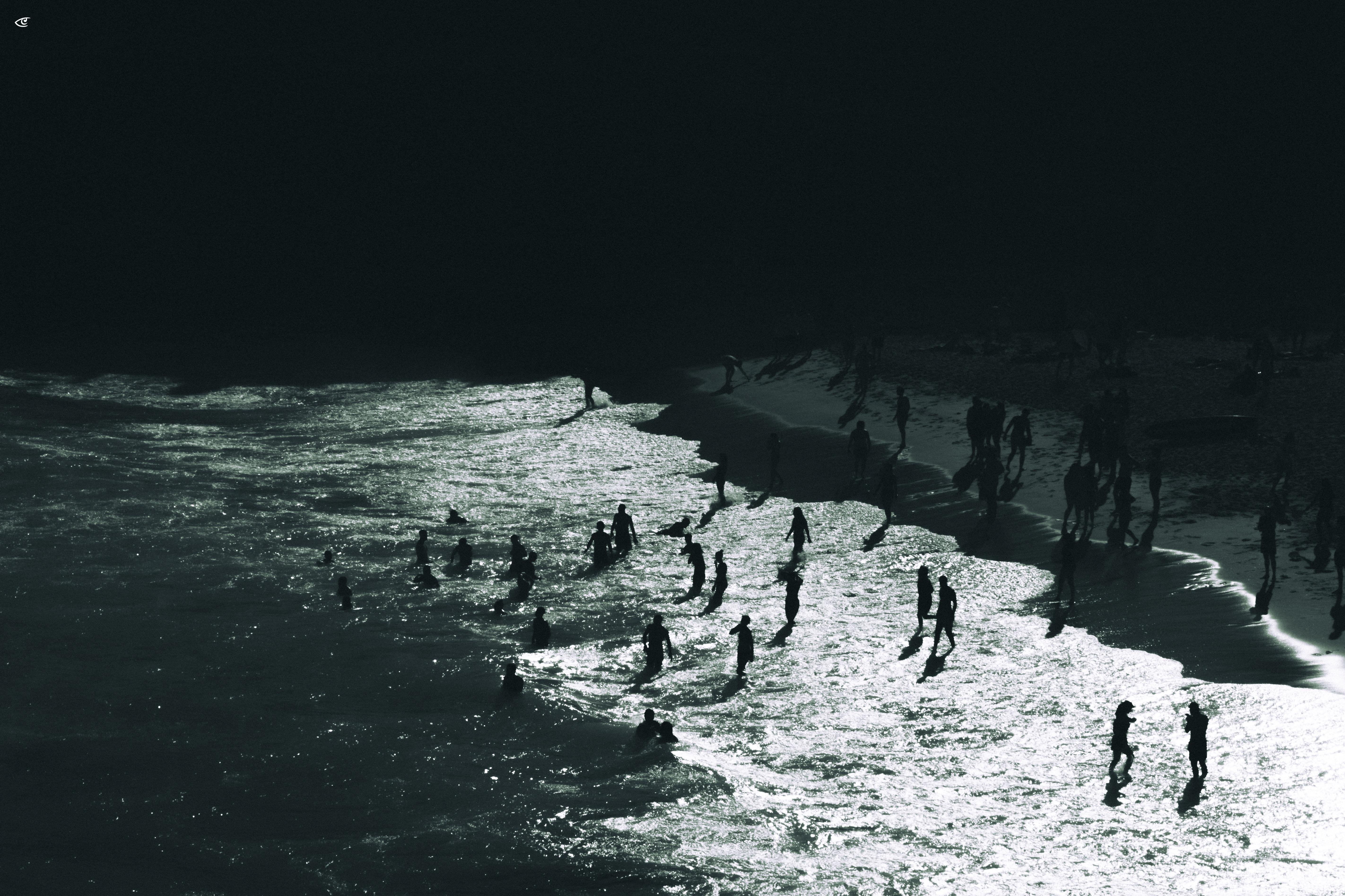 High-angle view of a beach shoreline with many people silhouetted in shallow surf; bright sunlit water along the lower right contrasts with a dark, shaded beach and background.
