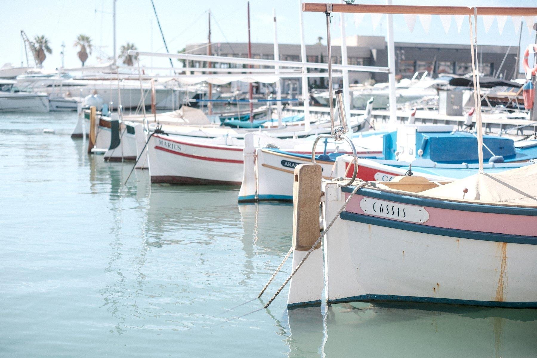 Photo horizontale. Une rangée de bateaux garés, tous blanc, le premier est avec une ligne rose bordé de bleu, avec une plaque blanche avec le nom de l’embarcation : « Cassis »