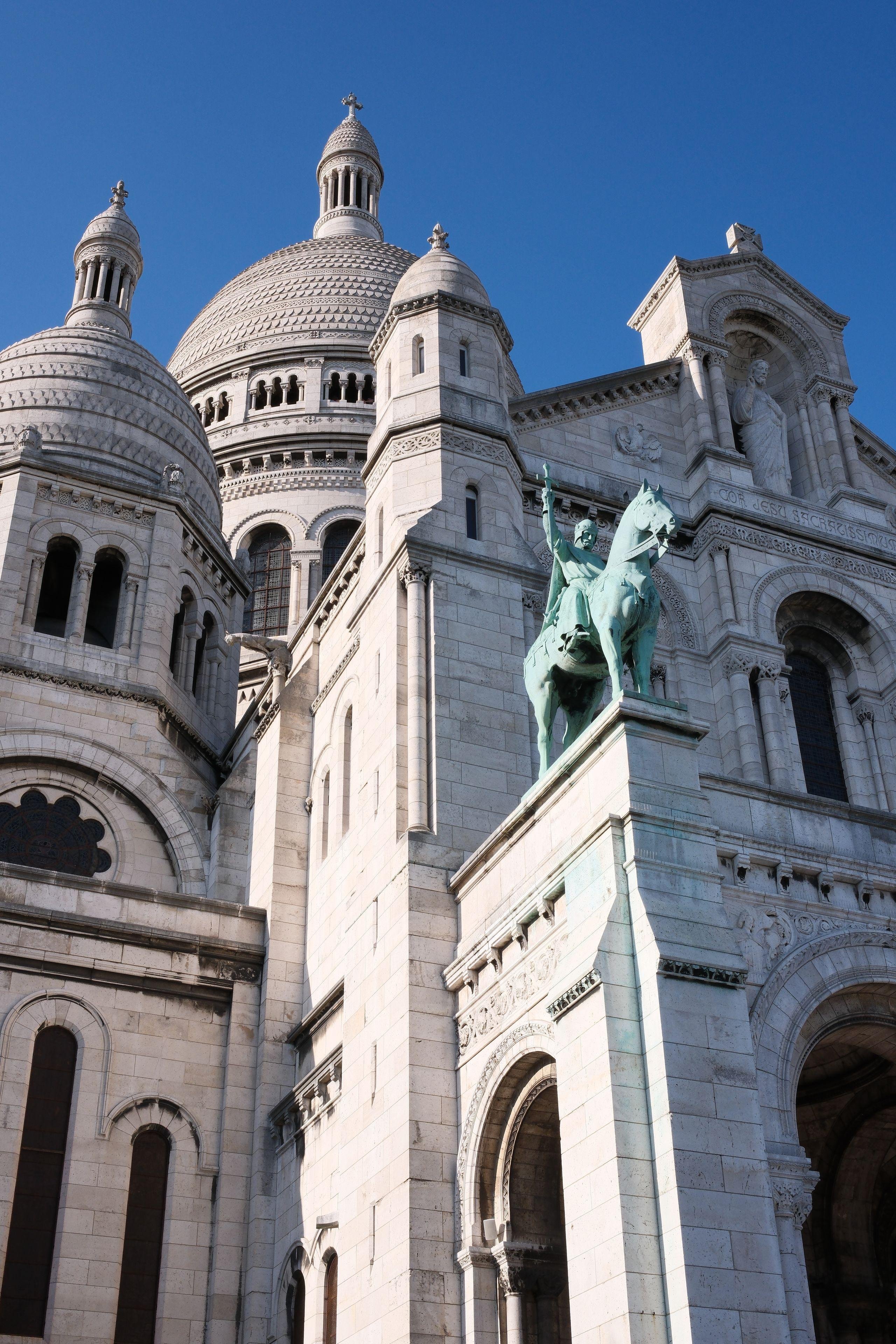 A low angled picture of the Sacré Cœur in Paris with a green statue of a green horse rider, which contrasts with the white of the church, under a bright blue sky in a very sunny day