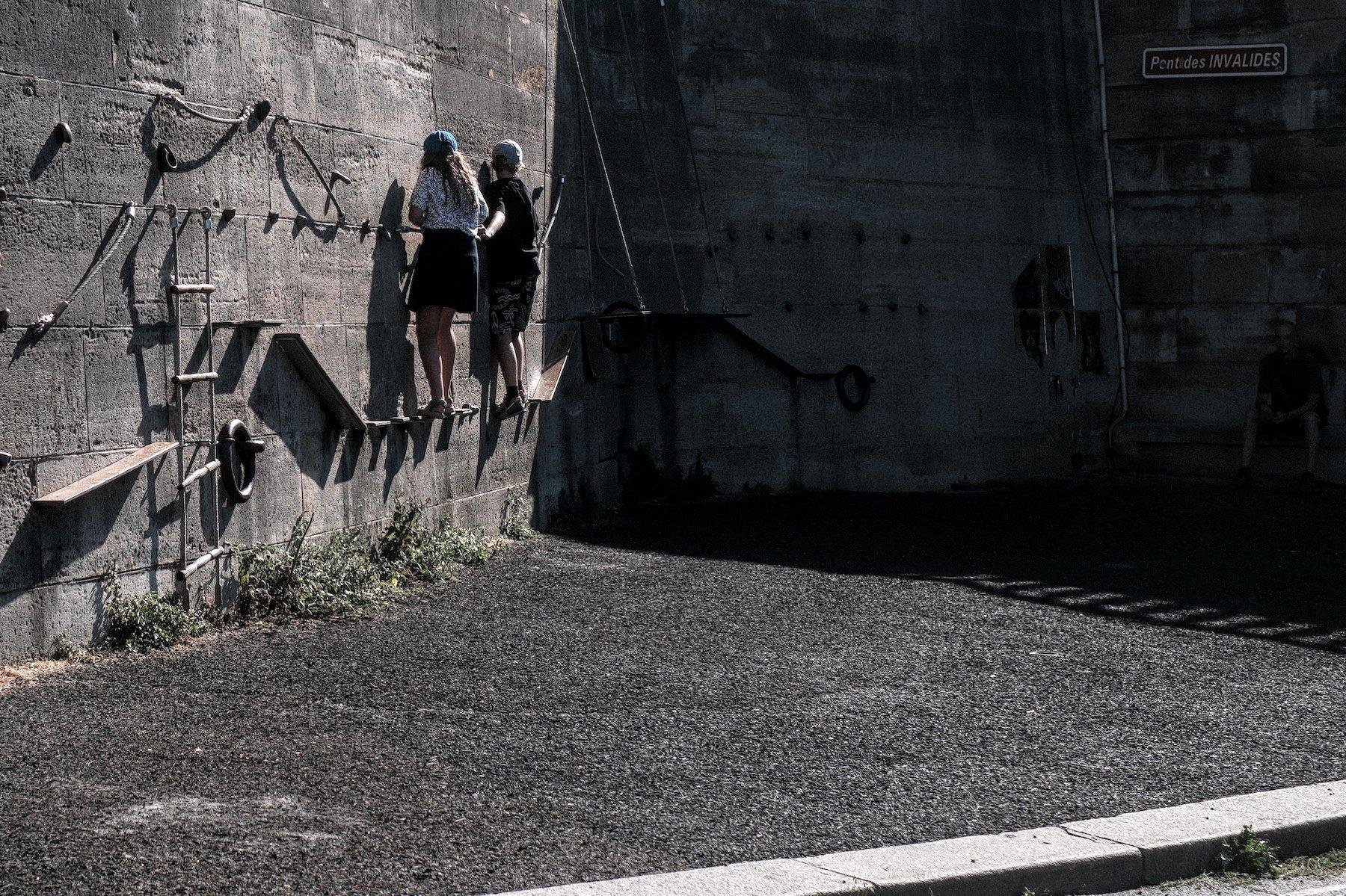 Photo horizontale. Sur un mur, des cordes épaisses, une échelle en corde et des prises d’escalades sont fixées. 2 enfants sont entrain de marcher sur une corde en tenant à une autre plus haut, sur le mur éclairé par le soleil couchant. Le mur change un peu d’angle par la suite, dans l’ombre