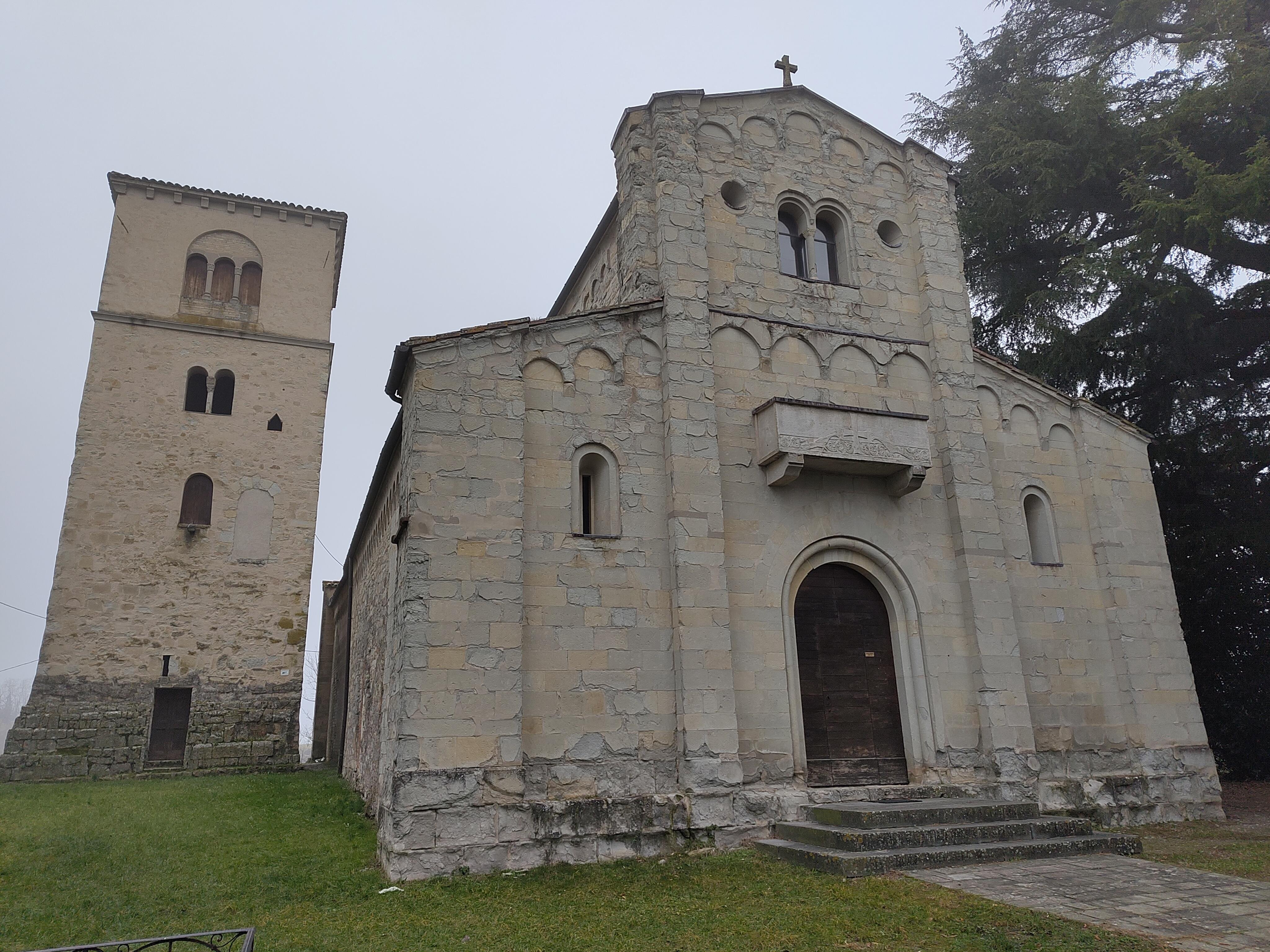 The image shows a small Romanesque-style church with a bell tower. The church has a simple stone facade with arched windows and a cross on top. The bell tower is adjacent and separate, featuring arched openings. The setting appears moderately rural, with a grassy area in the foreground and trees in the background.