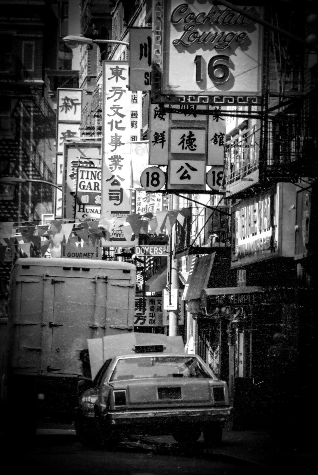 Crowded urban street scene in black and white featuring numerous vertical and horizontal signs with Chinese and English text. A vintage car and a large truck are visible in the foreground, partially obscured by the dense signage above. Overhead, strings of triangular flags hang between buildings. The signs advertise various businesses, including a cocktail lounge. Fire escapes are seen on building facades.