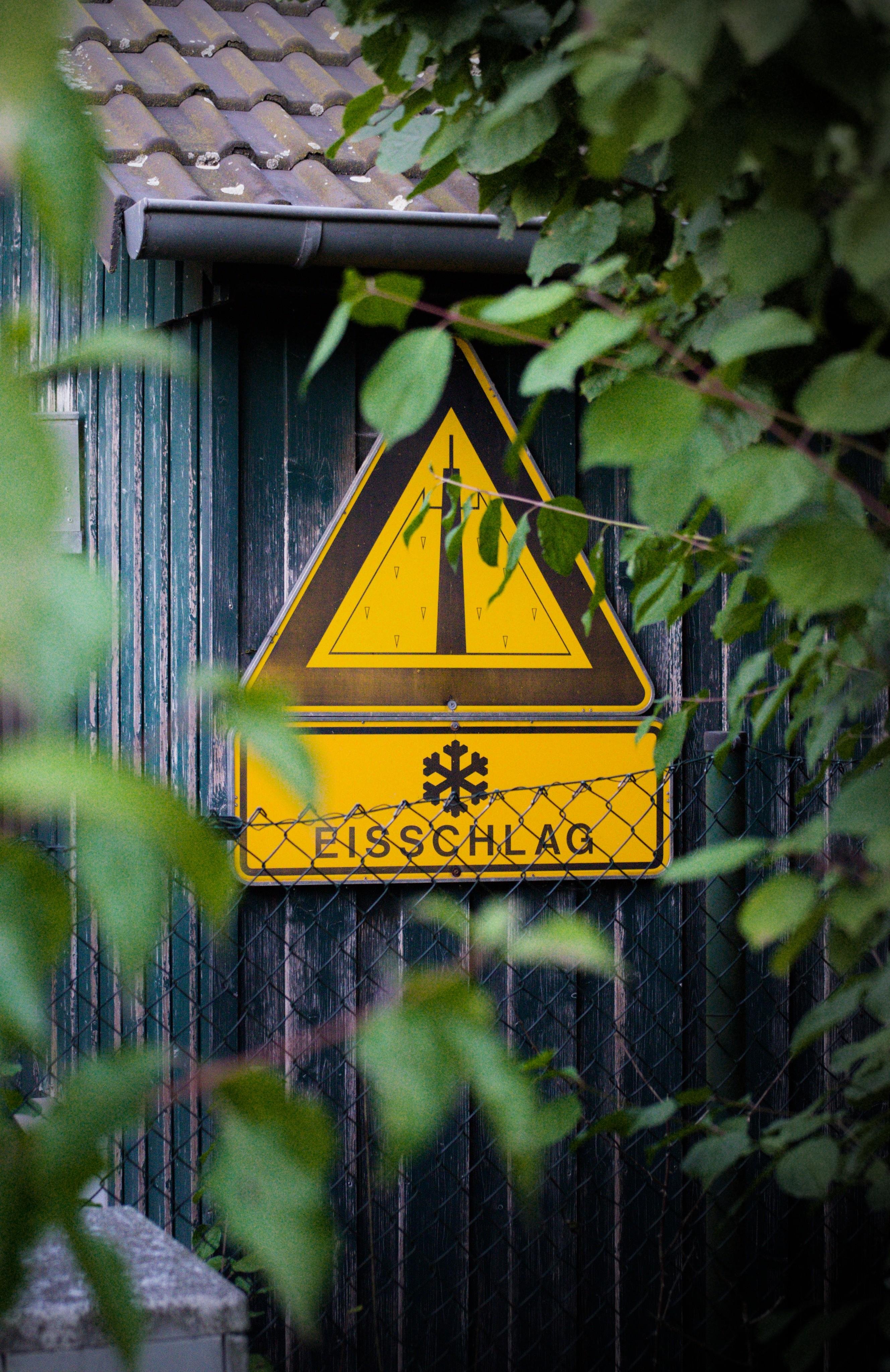 Yellow triangular warning sign with icicle graphic and a snowflake, labeled “EISSCHLAG,” mounted on a corrugated metal wall behind a chain-link fence, partially obscured by leafy branches in the foreground.