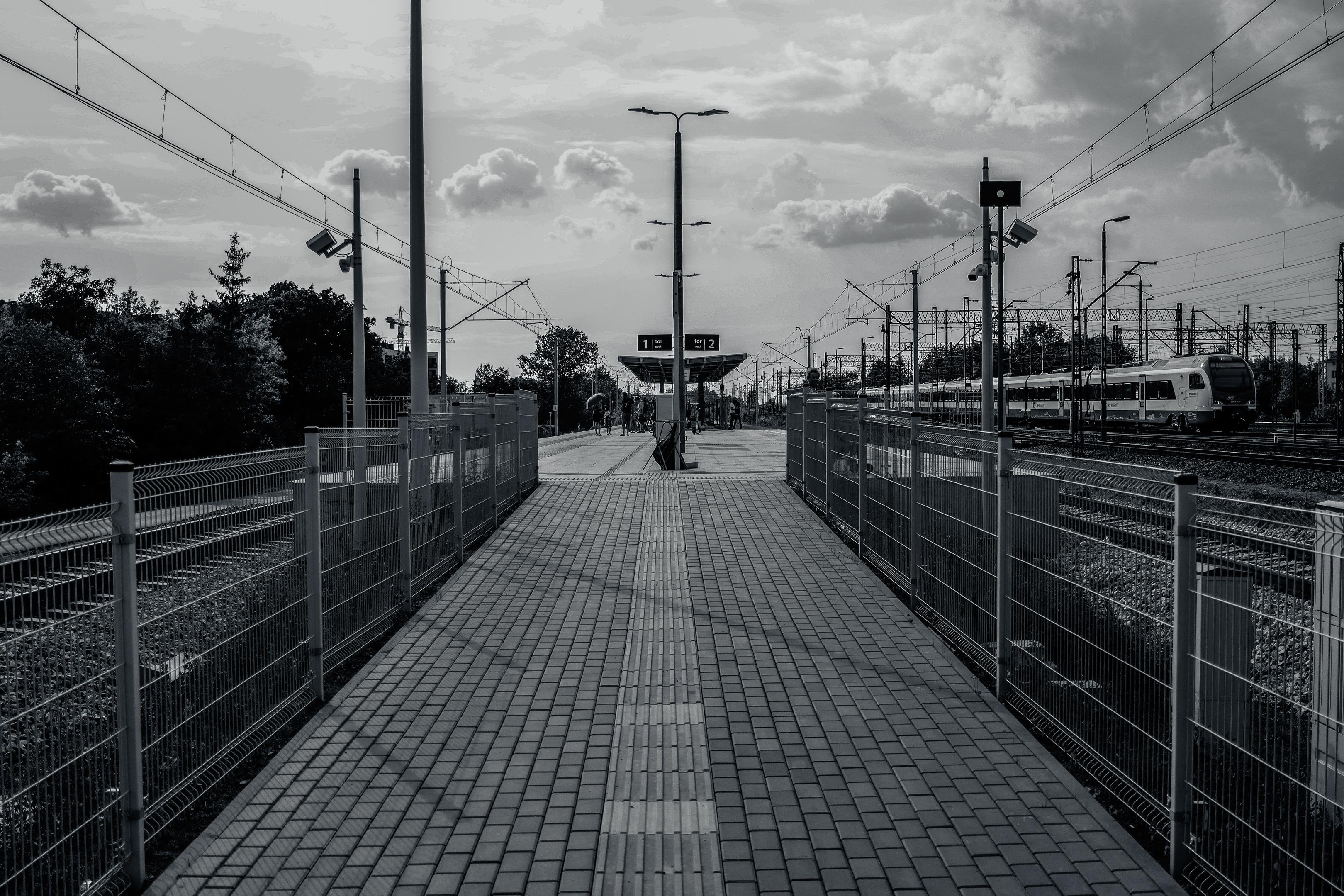 The image depicts a black-and-white photograph of a train station platform. It shows tracks to the right, with a train visible in the distance. The platform is lined with fences, and there are signs indicating platform numbers 1 and 2 in the center. The sky is cloudy, and there are overhead power lines for the trains. The overall scene is symmetrical and has an industrial feel.