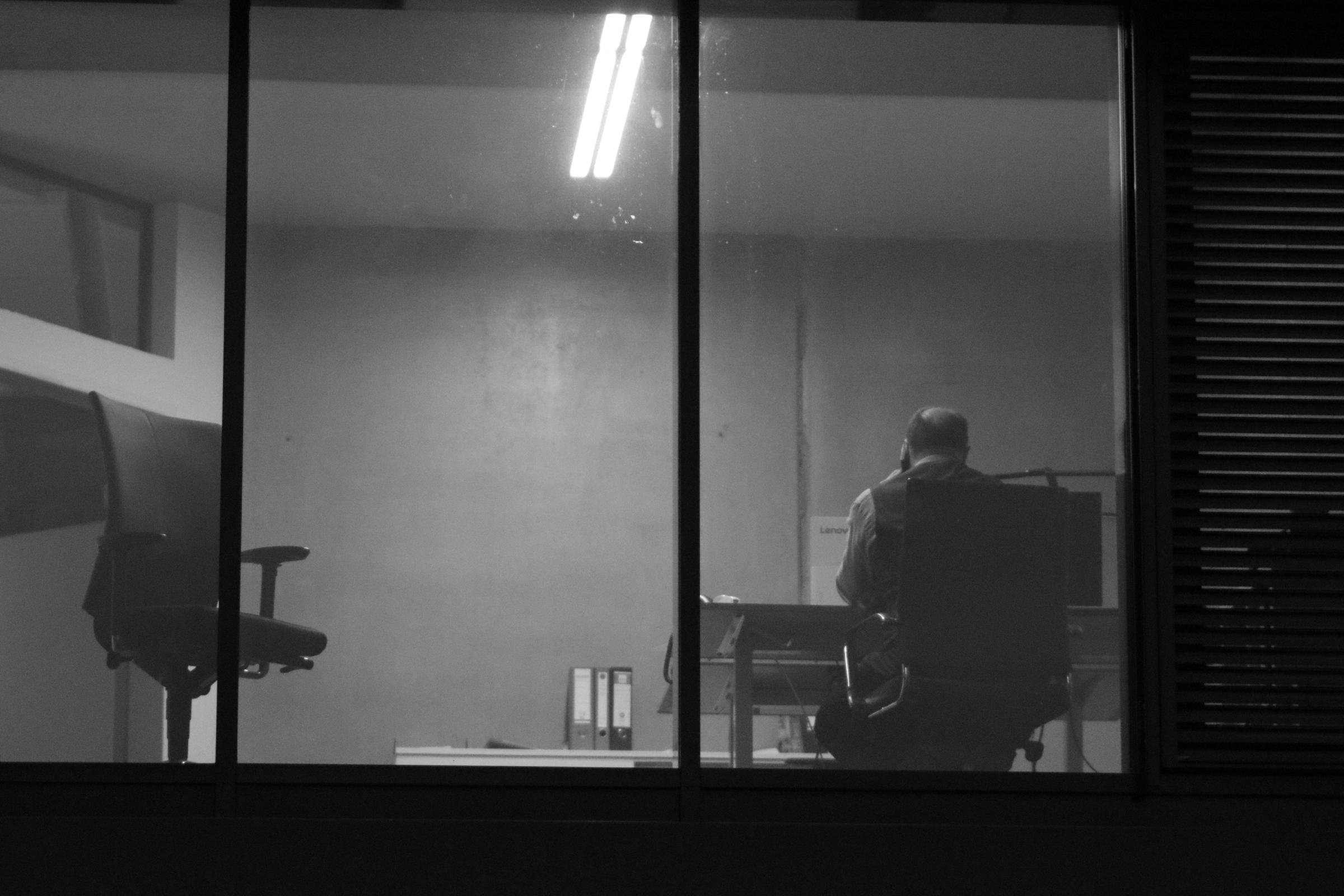 Black-and-white view through large office windows of a person seated at a desk facing away, with an empty office chair on the left, fluorescent ceiling lights above, binders on a shelf, and blinds along the right edge.

Shot with a cheap Benoison 85mm f/1.8 china-lense, which is much more better, than it i thought.