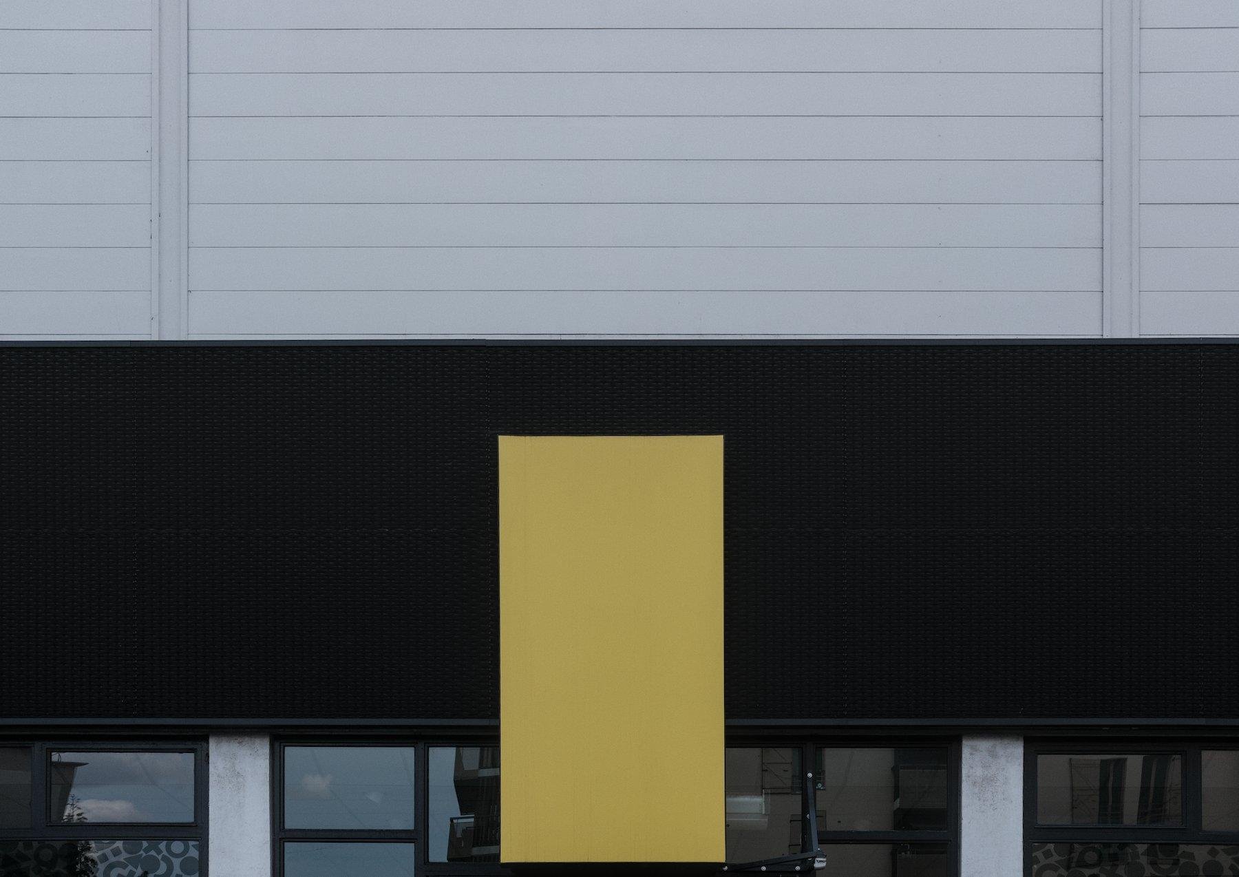 Photo horizontale. Une façade de bâtiment, faite de bandes grise en haut, d’un fond noir en dessous, des vitres encore en dessous, et devant le verre et le fond noir, un rectangle vertical jaune