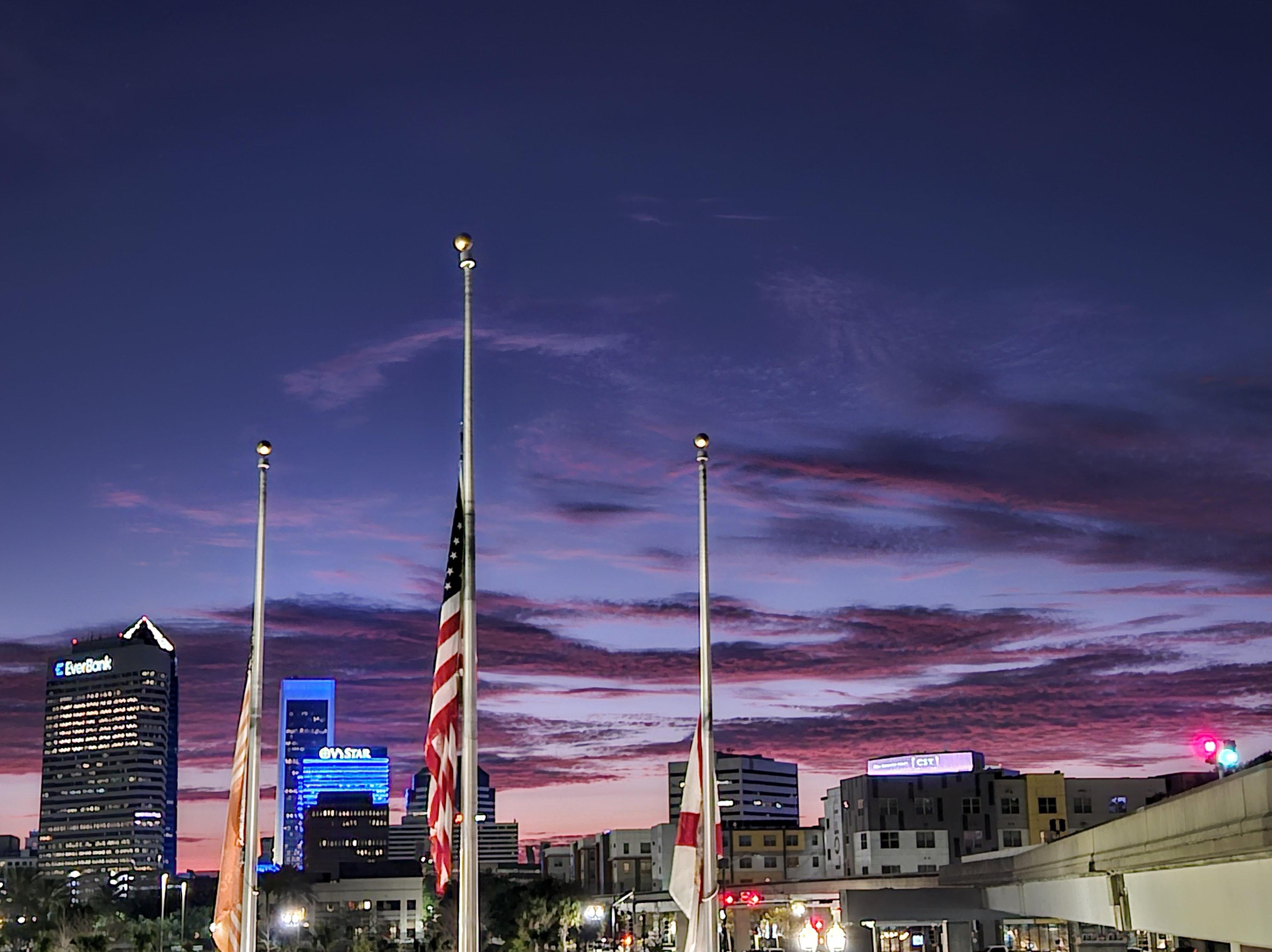 City skyline at twilight with a deep purple and pink sky. Three flags on tall poles in the foreground, one of which is the American flag. Several illuminated buildings with visible logos, including one for EverBank. A few smaller buildings and palm trees below against the colorful sky, creating an urban silhouette.