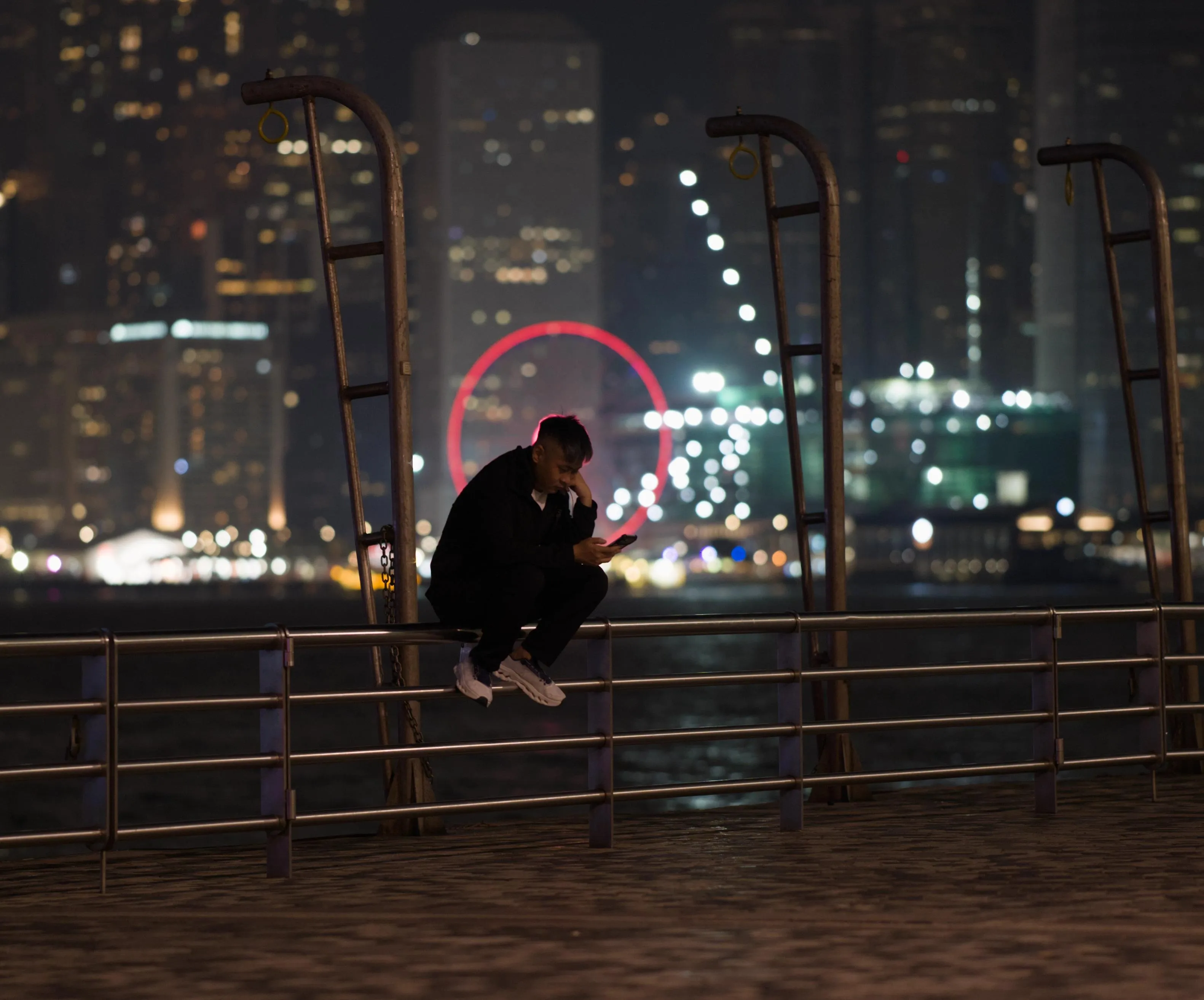 Nighttime photograph of a man sitting on the metal bars making up the railing of a waterside promenade. On the other side of the body of water, skyscrapers and city lights can be seen far away in the out-of-focus background blur. Some of these lights form a perfect circle with the head of the man at its center, looking like a red aureola.