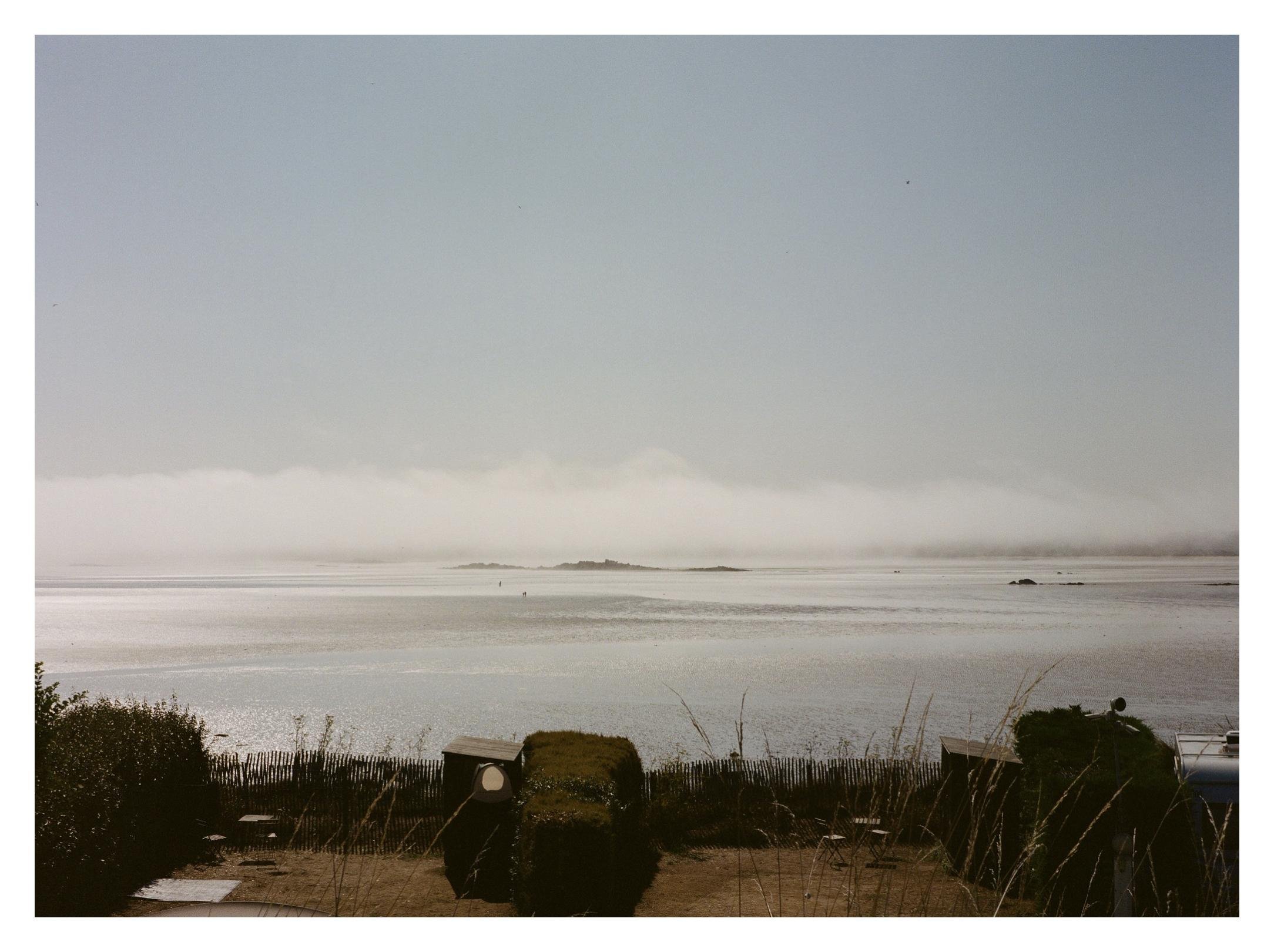 Wide view of a calm bay with small islands on the horizon under a gray sky, a low band of fog across the water, and coastal plants and a low fence in the foreground.