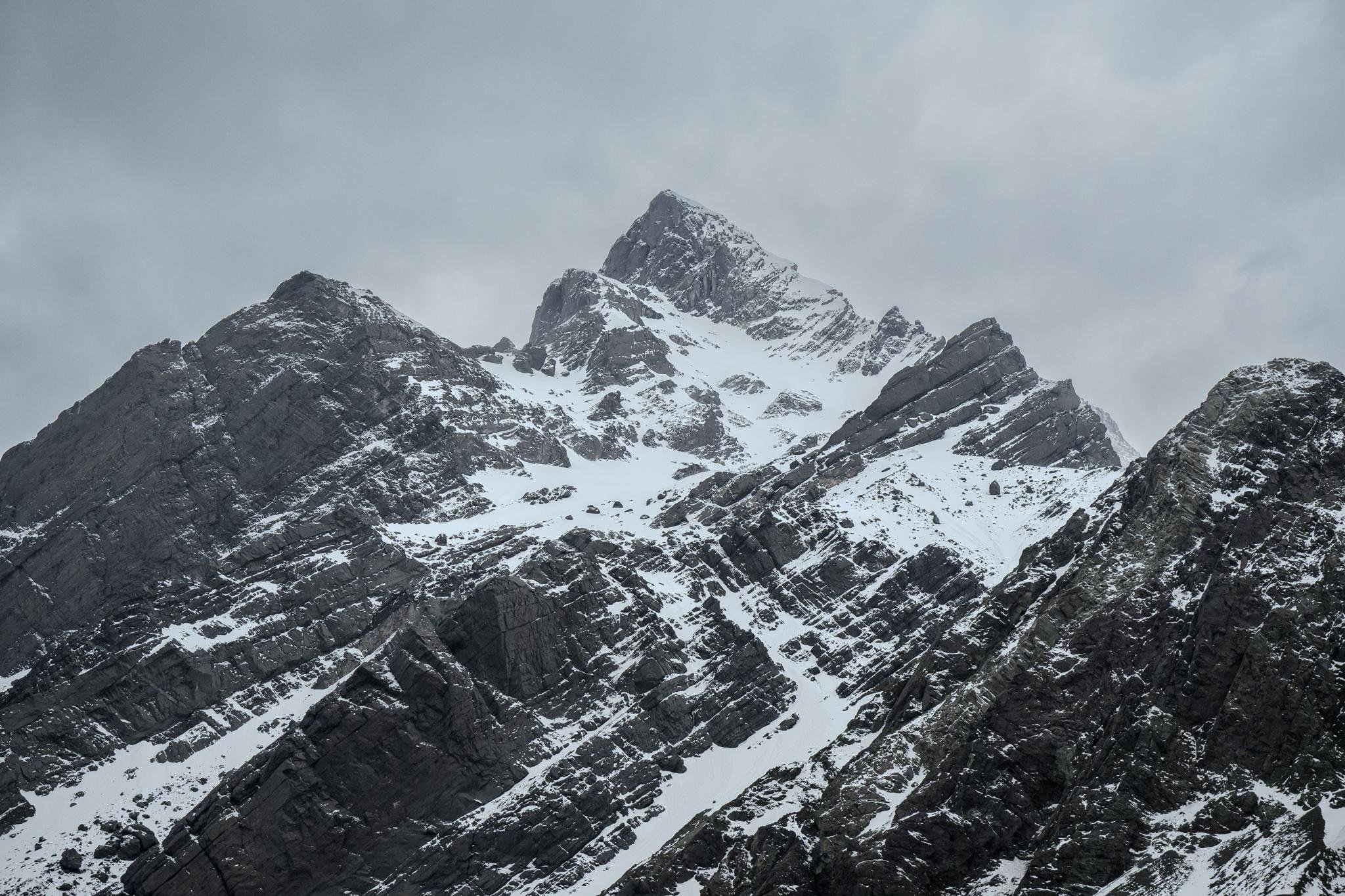 The image shows a rugged, snow-covered mountain (Aconcagua) under an overcast, cloudy sky. The slopes are steep, with rocky outcrops visible among the snow.