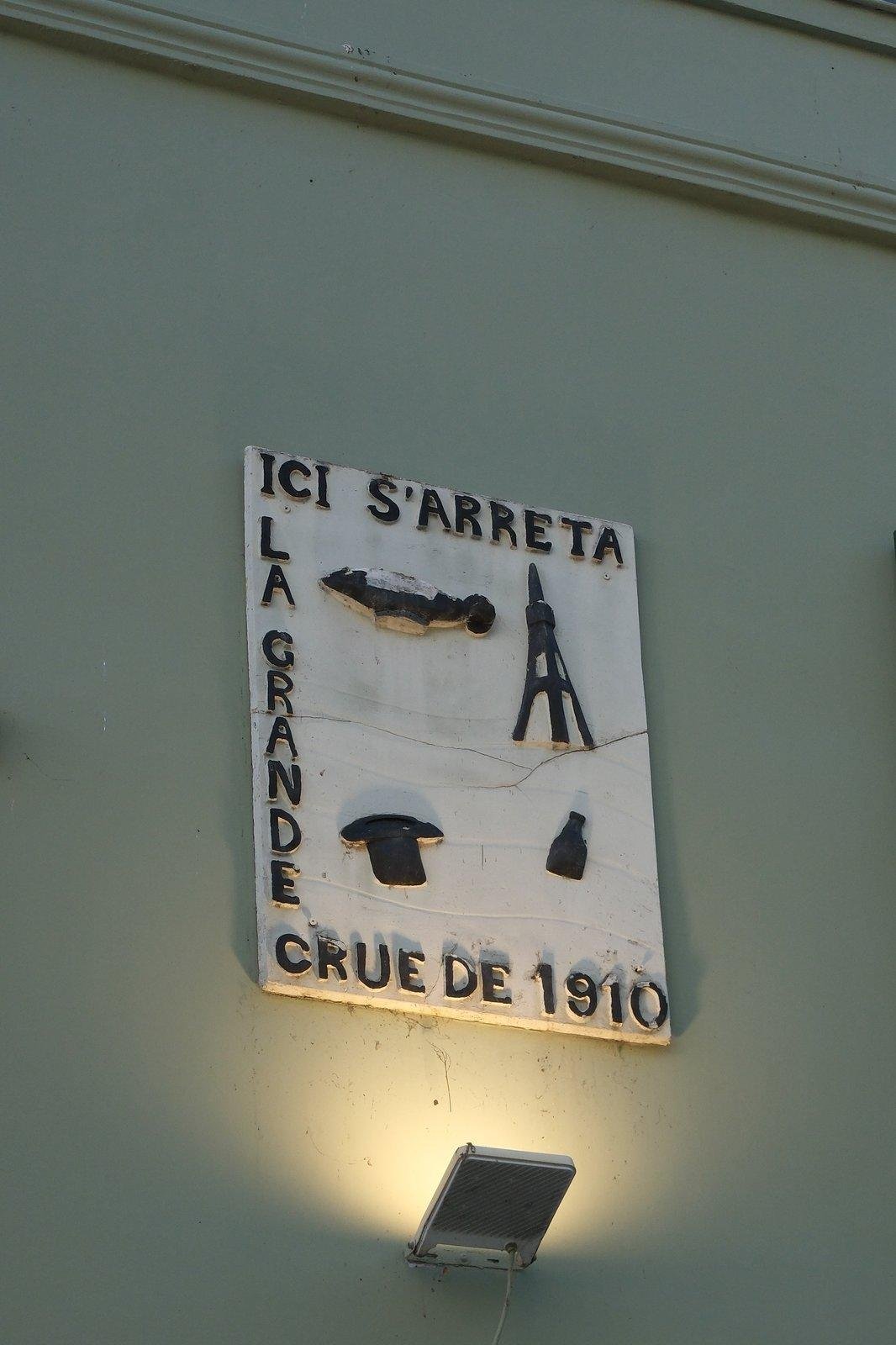 Une petite plaque commémorant la crue de la Seine de 1910 avec une tour Eiffel stylisée, un chapeau haut de forme à l’envers et une bouteille flottants dans l’eau et un truc qui ressemble à un poisson mais je ne crois pas que ce soit ça. On peut y lire : «&nbsp;Ici s’arrêta la grande crue de 1910&nbsp;»