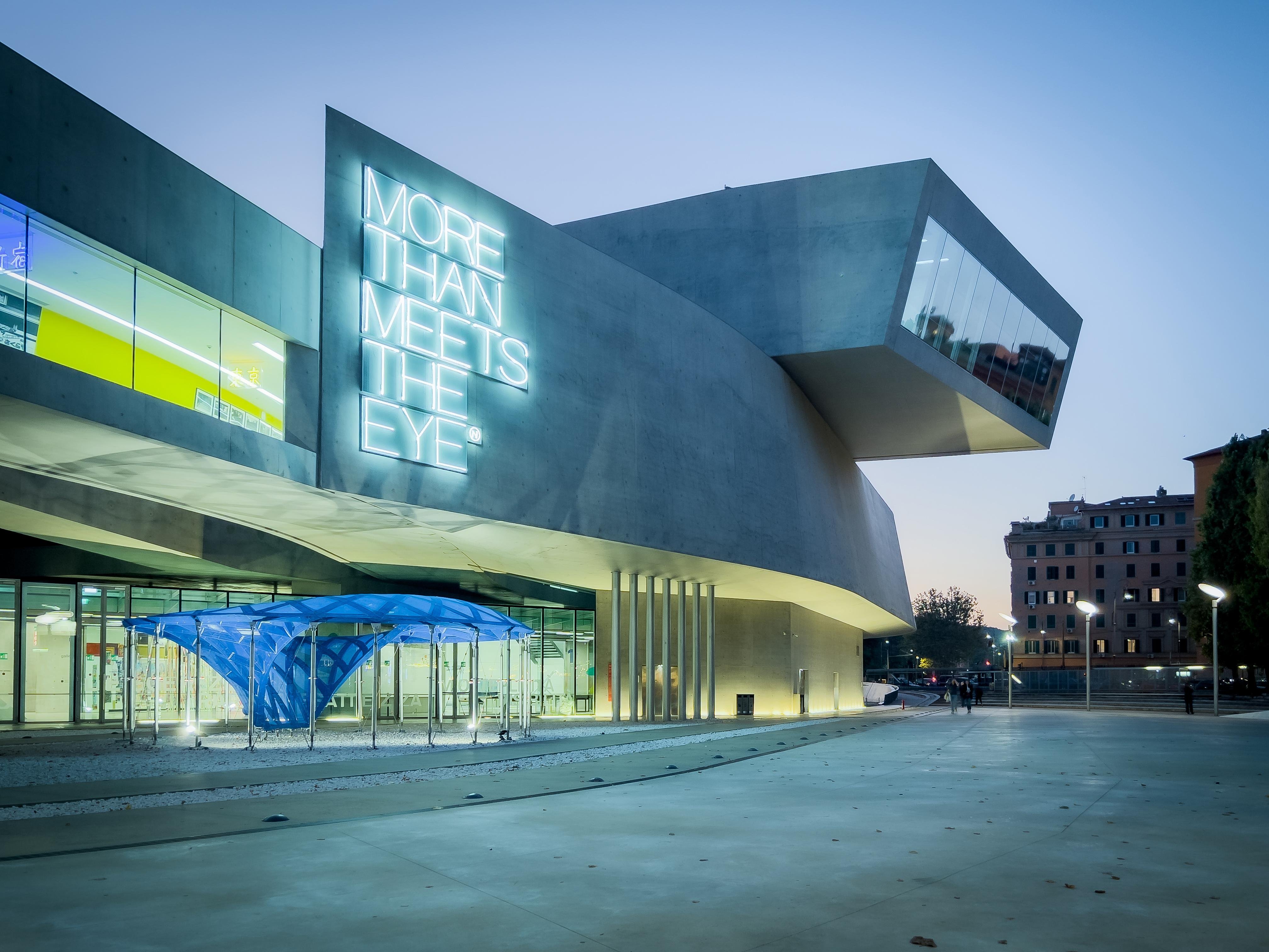 Edificio architettonico moderno con un design squadrato e sinuoso. C’è un’insegna al neon prominente sull’edificio che recita “MORE THAN MEETS THE EYE”. Una struttura con un tendone blu è situata davanti all’edificio. La scena è ambientata all’alba o al tramonto, con un’illuminazione soffusa che esalta le caratteristiche dell’edificio