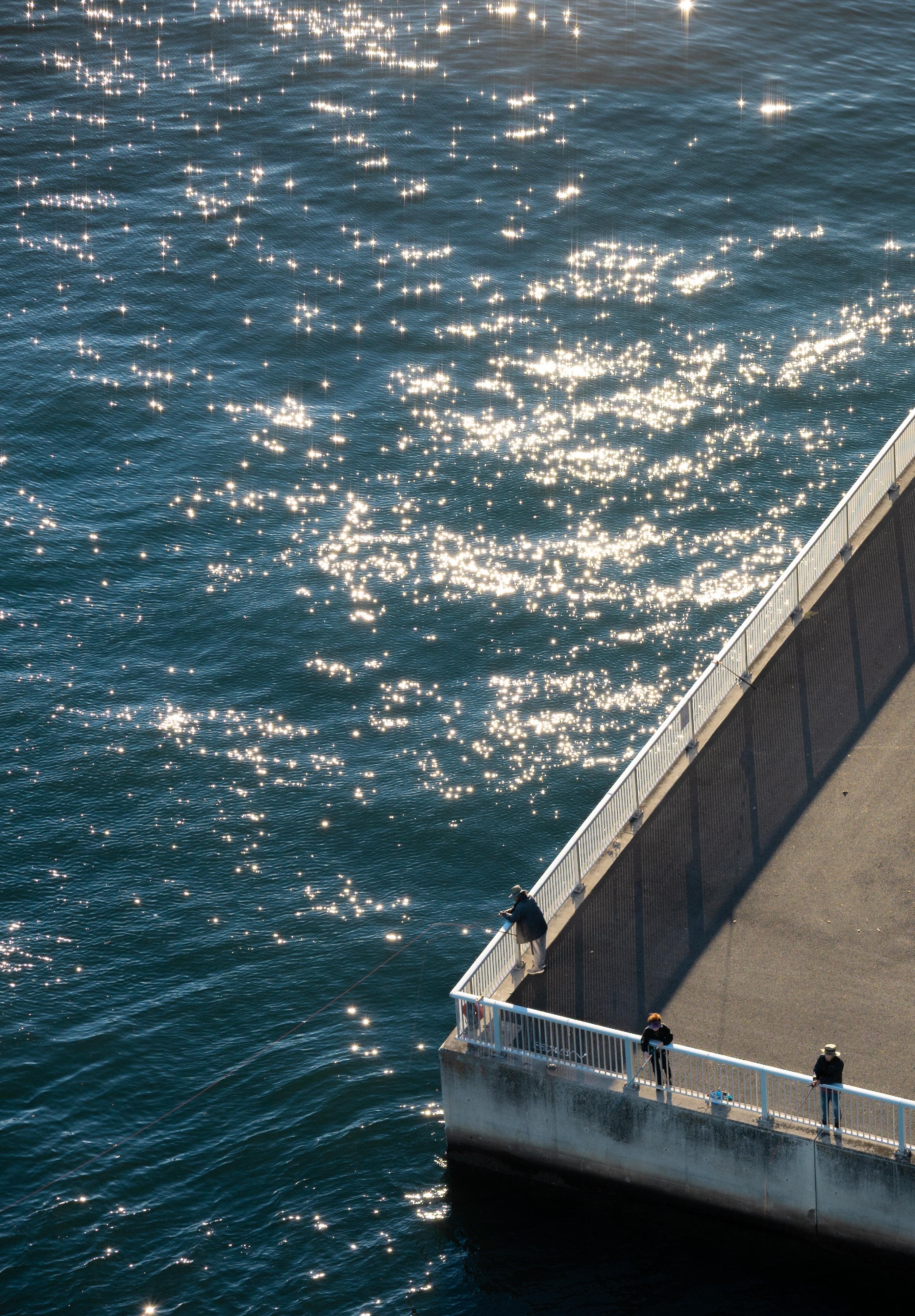 Aerial view of a waterfront with a concrete walkway bordered by a metal railing. Sparkling sunlight reflects on the water's surface. Several people are positioned along the walkway, some engaged in fishing. A bicycle is parked near the grassy area with neatly arranged steps and a lamppost, casting elongated shadows onto the ground.