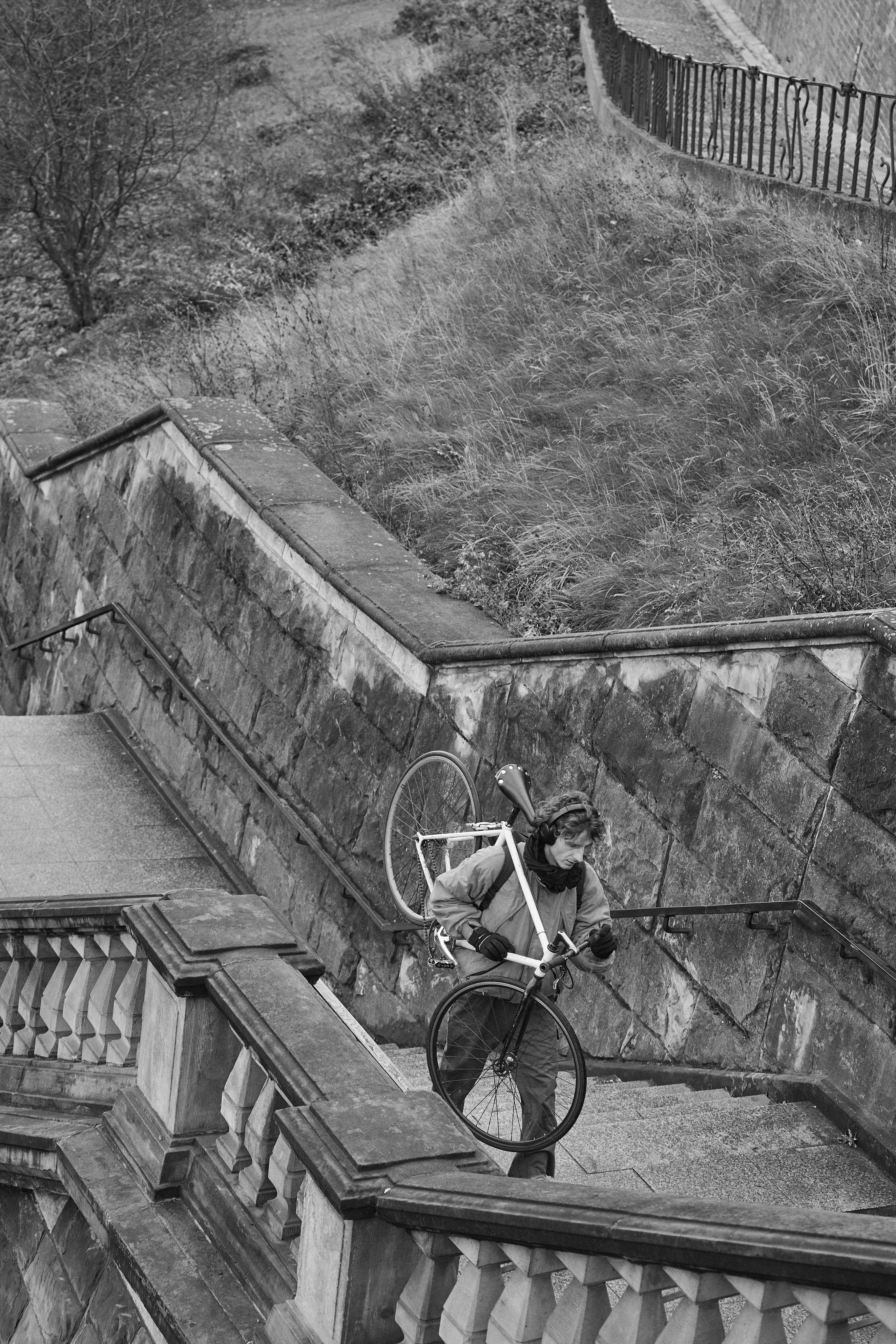 The image shows a person carrying a bicycle up a stone staircase. The setting appears to be outdoors with grass and a railing visible in the background. It's in black and white.