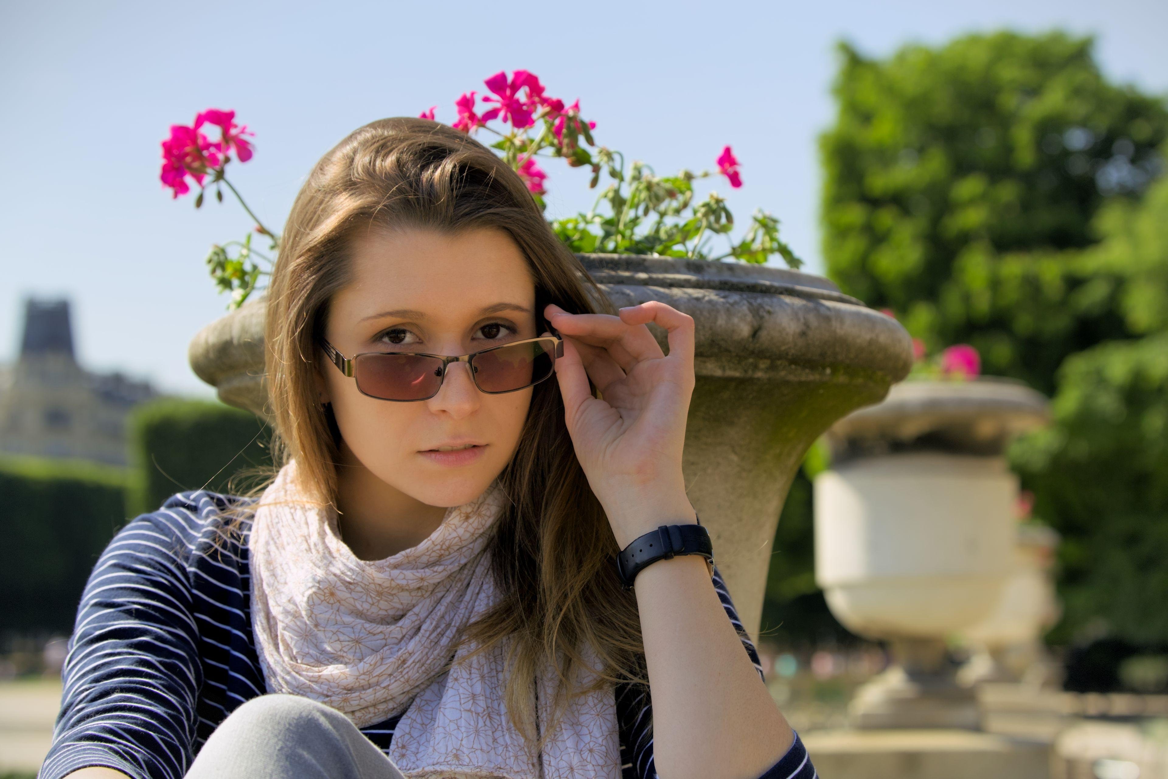 Photo format paysage d’une brune qui baisse ses lunettes de soleil de la main gauche pour regarder par dessus vers la caméra. Derrière elle se trouvent de gros pots de fleurs et des arbres