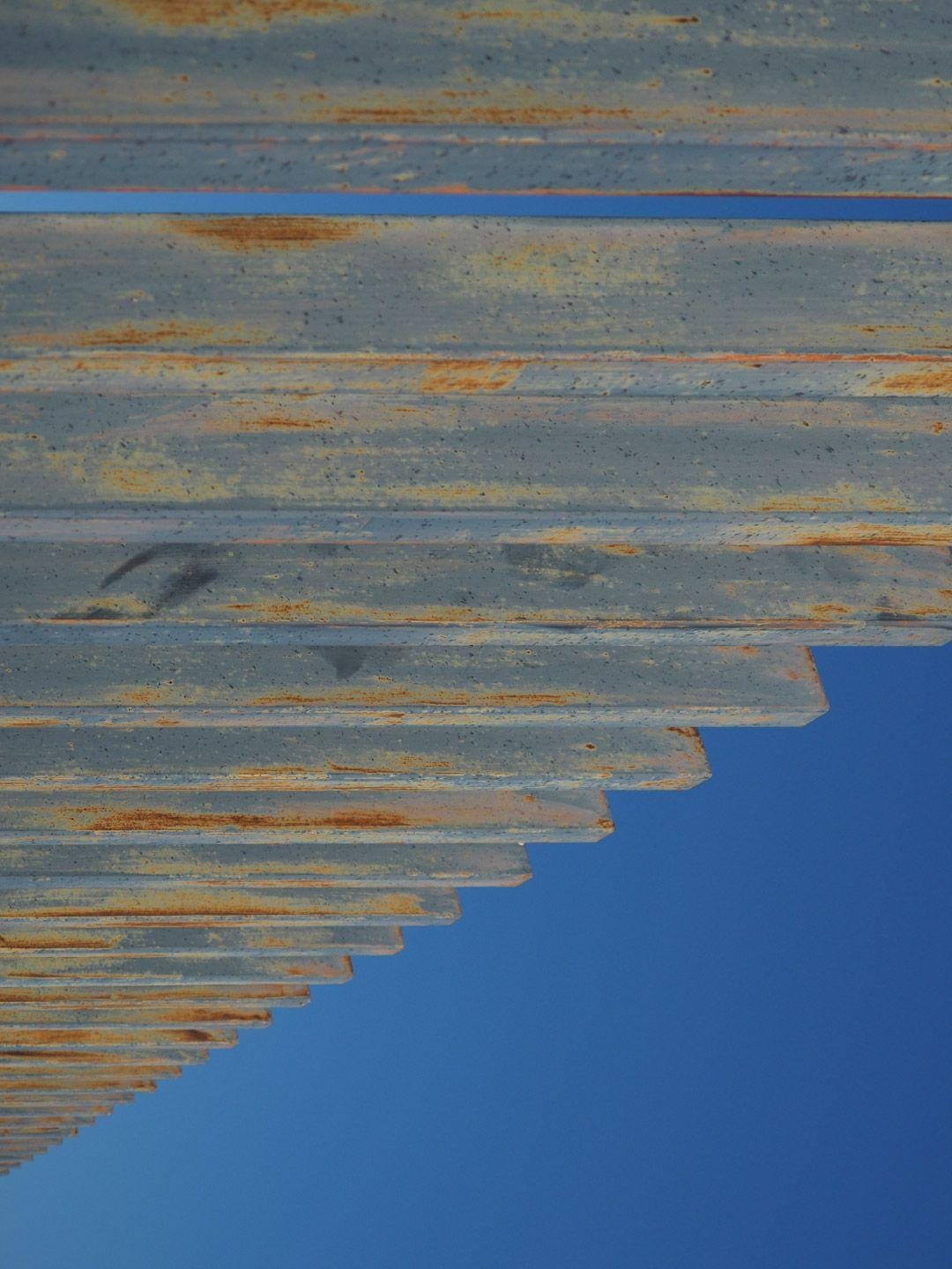 Elementi verticali di una staccionata metallica, con segni di ruggine, contro un cielo azzurro intenso. Foto ruotata di 90 gradi.