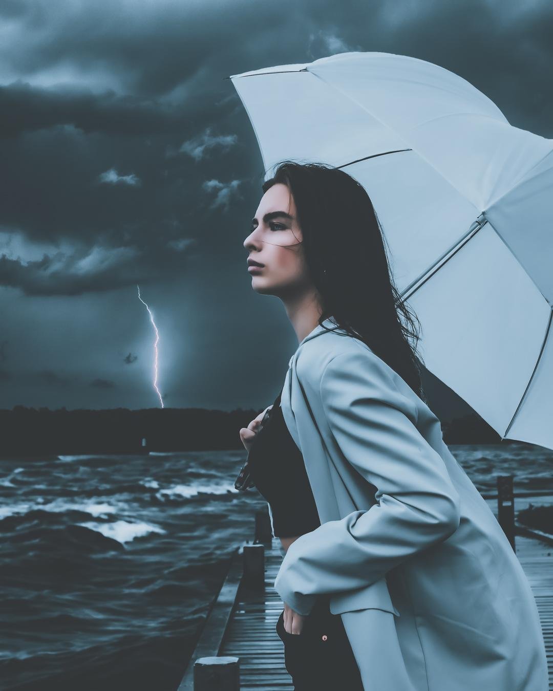 The image shows a person standing on a dock by a body of water during a storm. They are holding a light-colored umbrella, and there's a bolt of lightning visible in the sky behind them. The atmosphere is dramatic, with dark clouds and waves in the water. Valokuvaaja Kuvaajankulma Lappeenranta