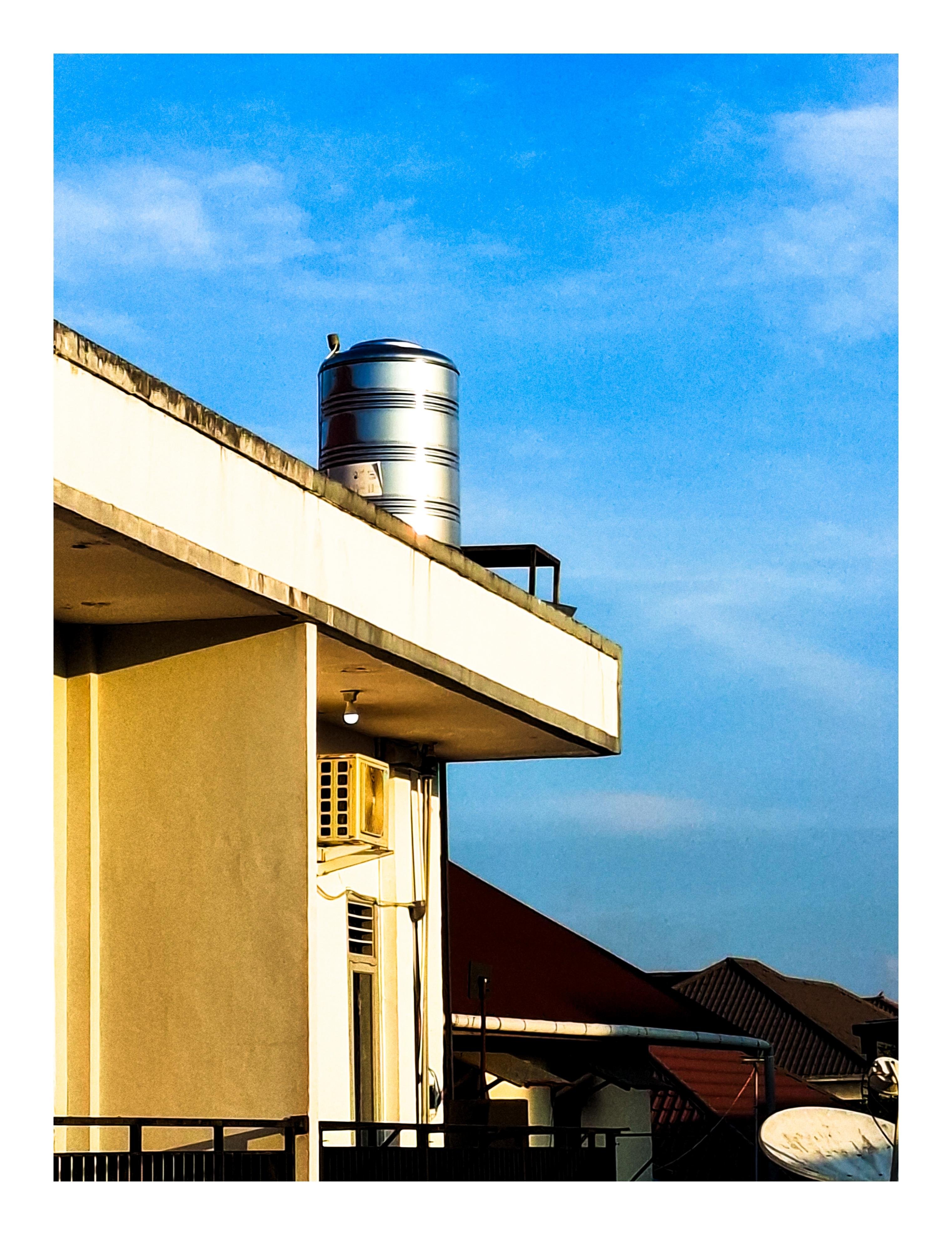 Corner of a flat-roofed building with a metal cylindrical water tank on the roof, an exterior air-conditioning unit on the wall, and a railing in the foreground; neighboring rooftops and a satellite dish below against a blue sky.