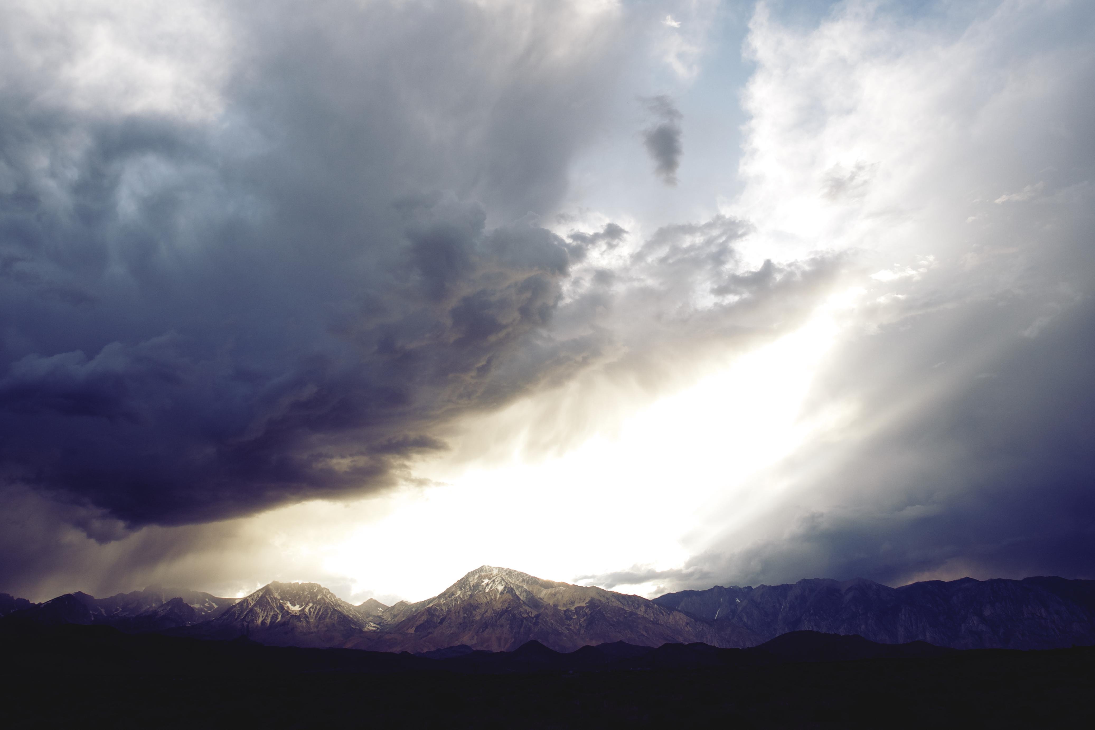 Dunkle Sturmwolken über einer fernen Bergkette, mit einem hellen Bruch in den Wolken, die Sonnenstrahlen auf schneebestaubte Gipfel über einem dunklen Vordergrundkamm werfen.