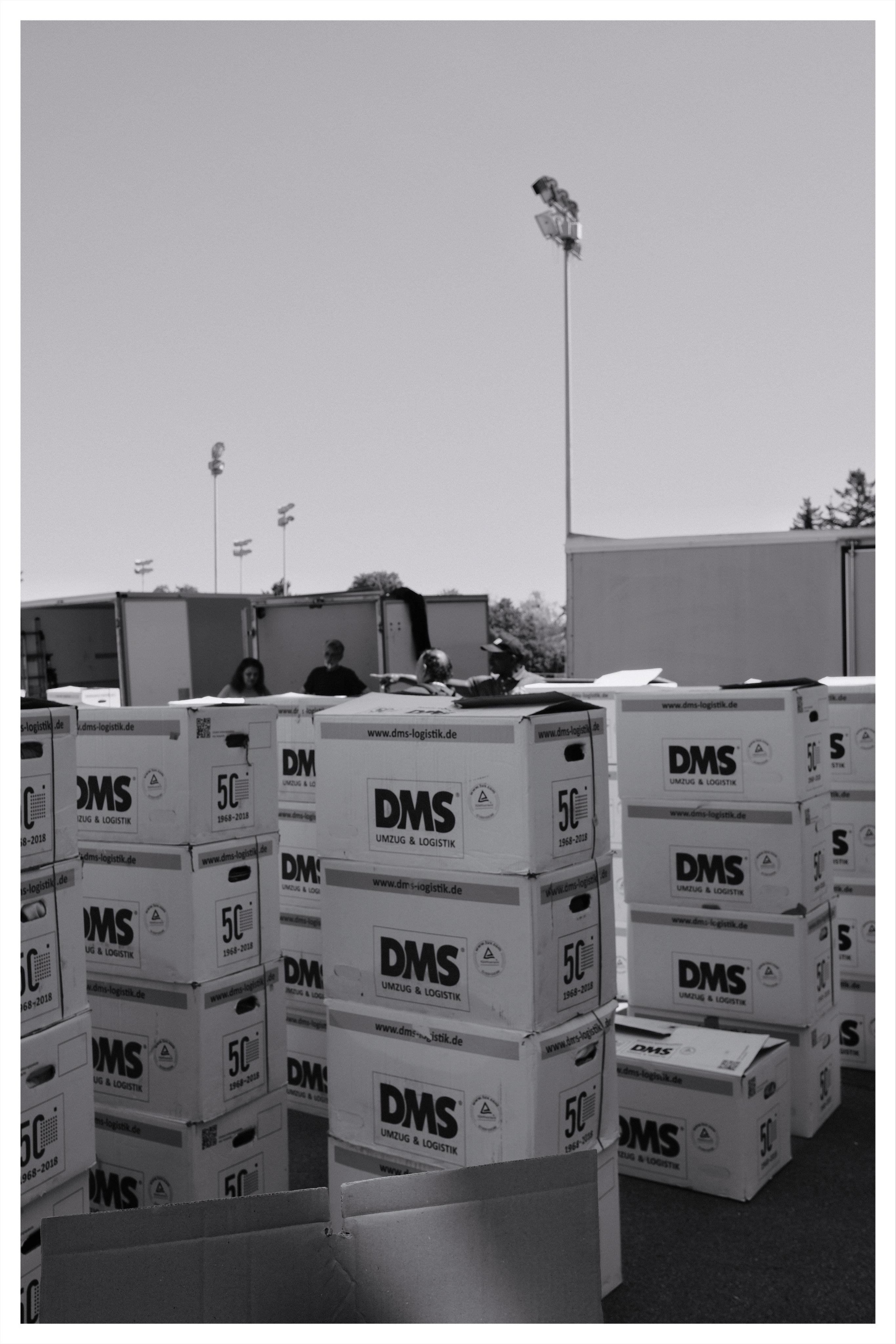 Stacks of cardboard moving boxes labeled “DMS Umzug & Logistik” outdoors, with open truck trailers and a few workers partially visible behind the boxes; tall light poles rise in the background under a clear sky.