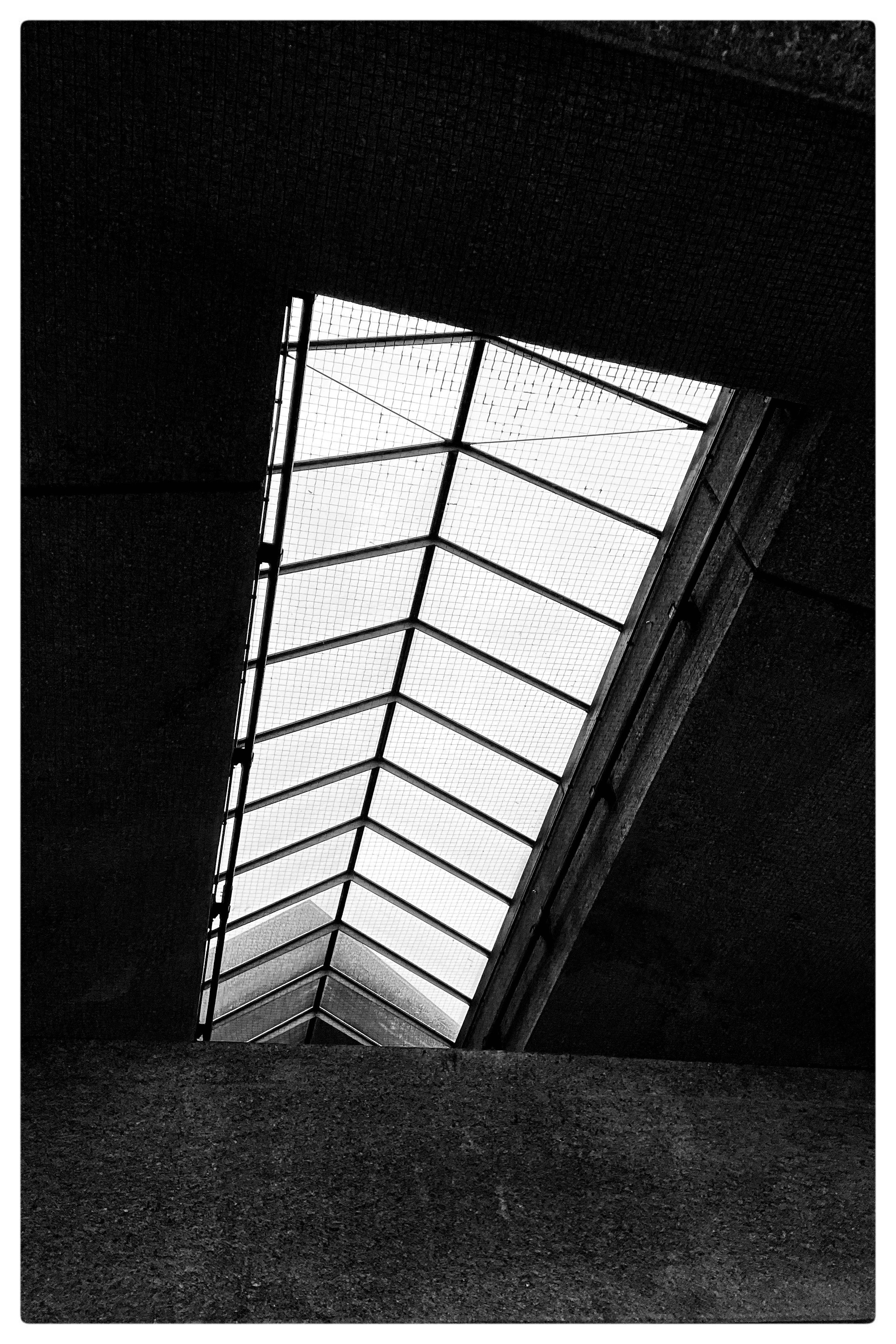 Triangular skylight with a grid pattern, framed by dark concrete beams. Black and white composition highlighting geometric lines and contrast between light roof and dark ceiling.