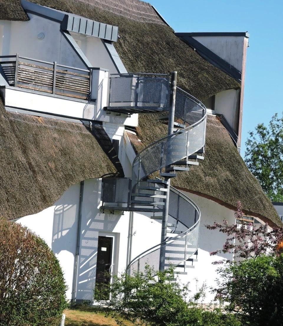 Spiral staircase attached to the exterior of a white building with a thatched roof. Metal railings border the staircase steps. Large windows and a small doorway are present on the building's facade. Green shrubs and trees surround the structure, and the sky is clear and blue.