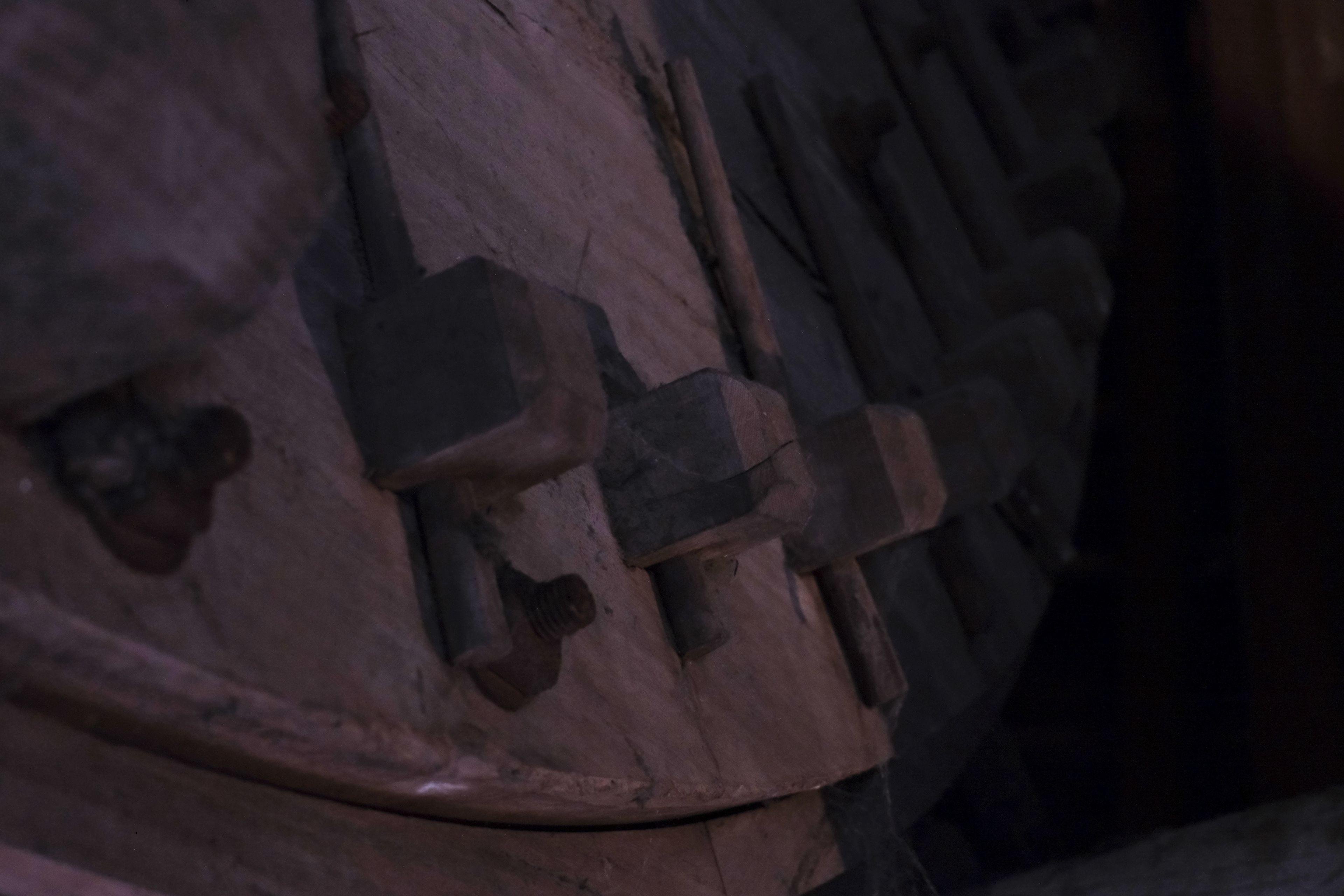 Photo format paysage montrant une partie de rouage de moulin à vent. On y voit un bout de disque massif en bois, surmonté à sa surface et probablement sur tout le périmètre, des cubes en bois dont les arrêtes supérieures ont été biseautées. De courtes tiges en métal traverse ces cubes en suivant le rayon, sur la surface du disque.