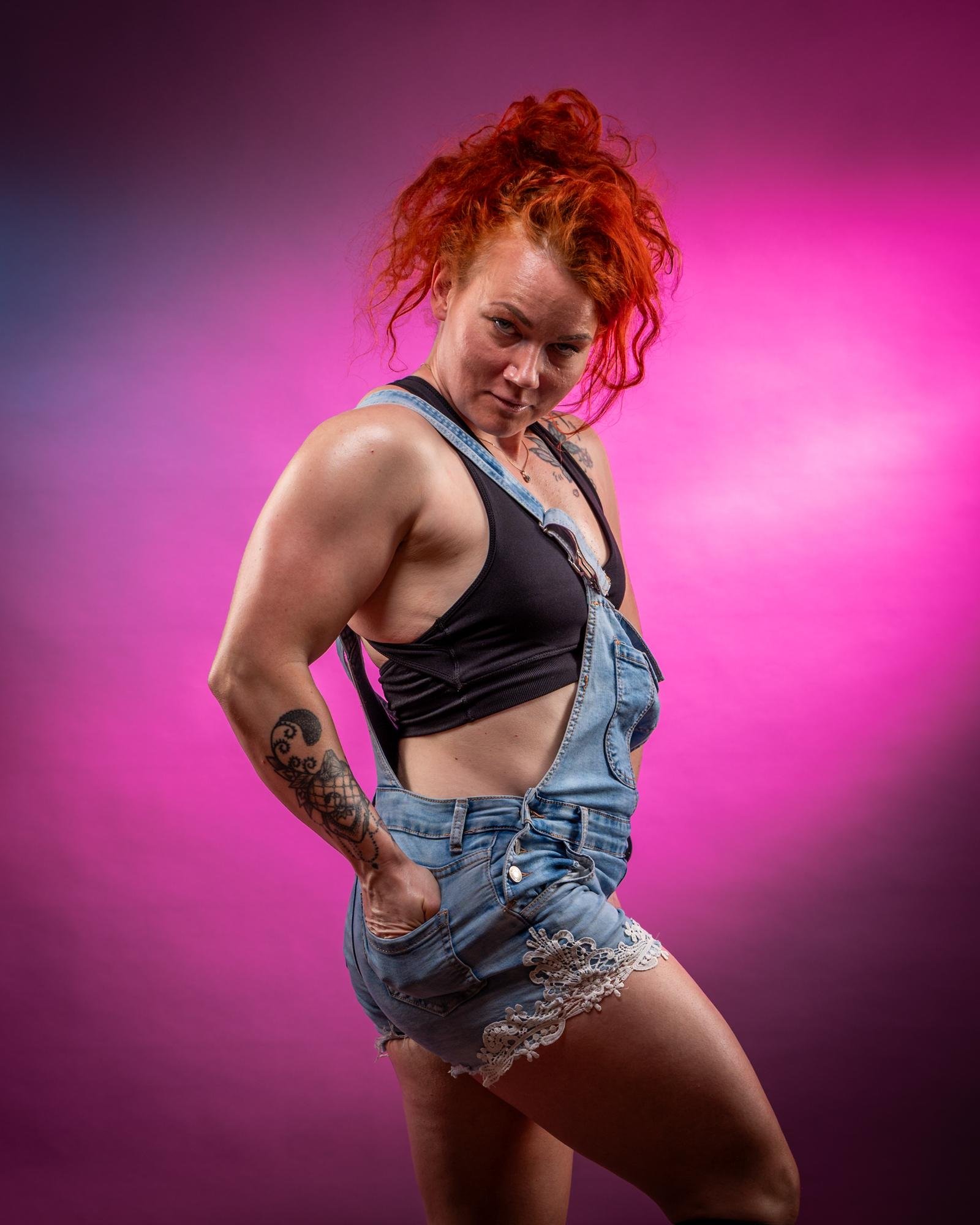 The image shows a person with red hair posing against a background with purple and pink tones. They are wearing denim overalls with lace trim, a black top, and they have tattoos on their arm. The style is dynamic and expressive.
