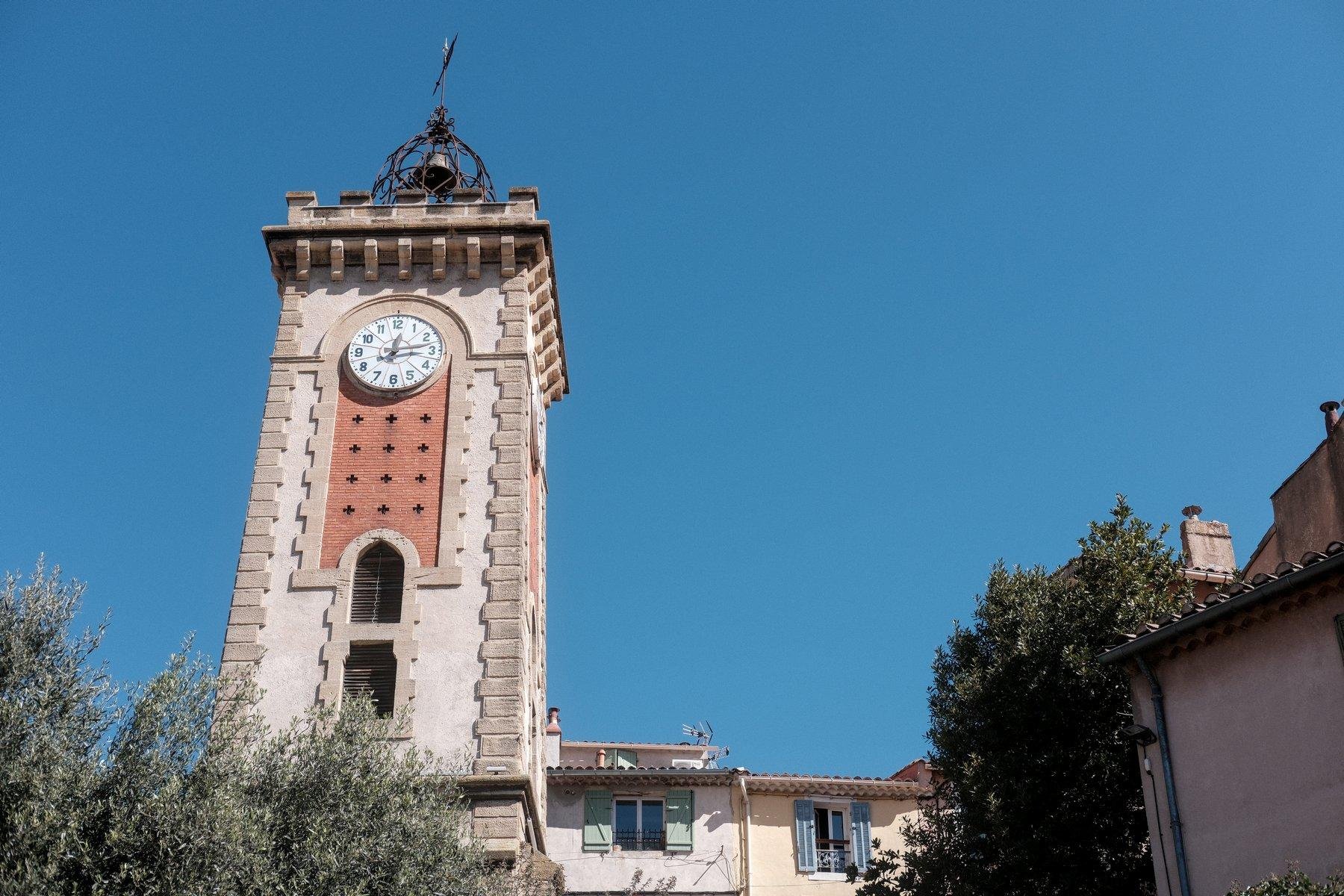 Photo horizontale. Contre-plongée sur le clocher d’Aubagne, une grande tour carré, avec une horloge, et au sommet, une cloche