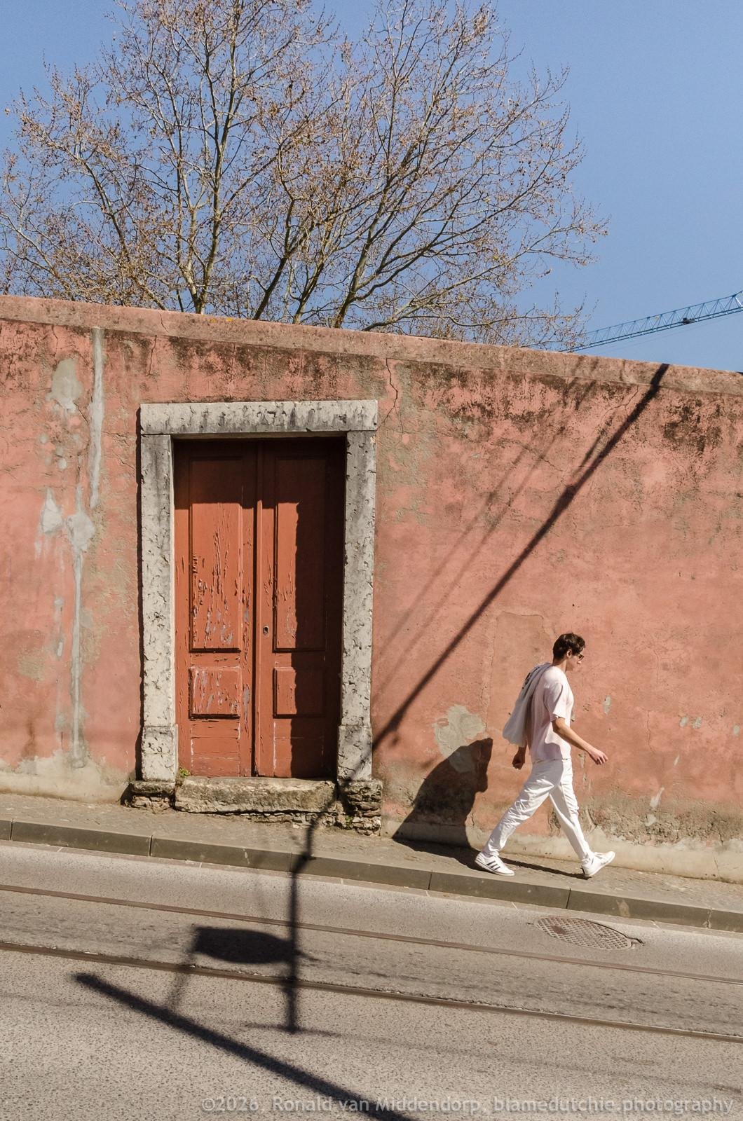 Person in light clothing walking along a sidewalk beside a weathered pink stucco wall with a faded red double door in a stone frame; bare tree branches and a crane rise above the wall, and strong diagonal shadows fall across the street.